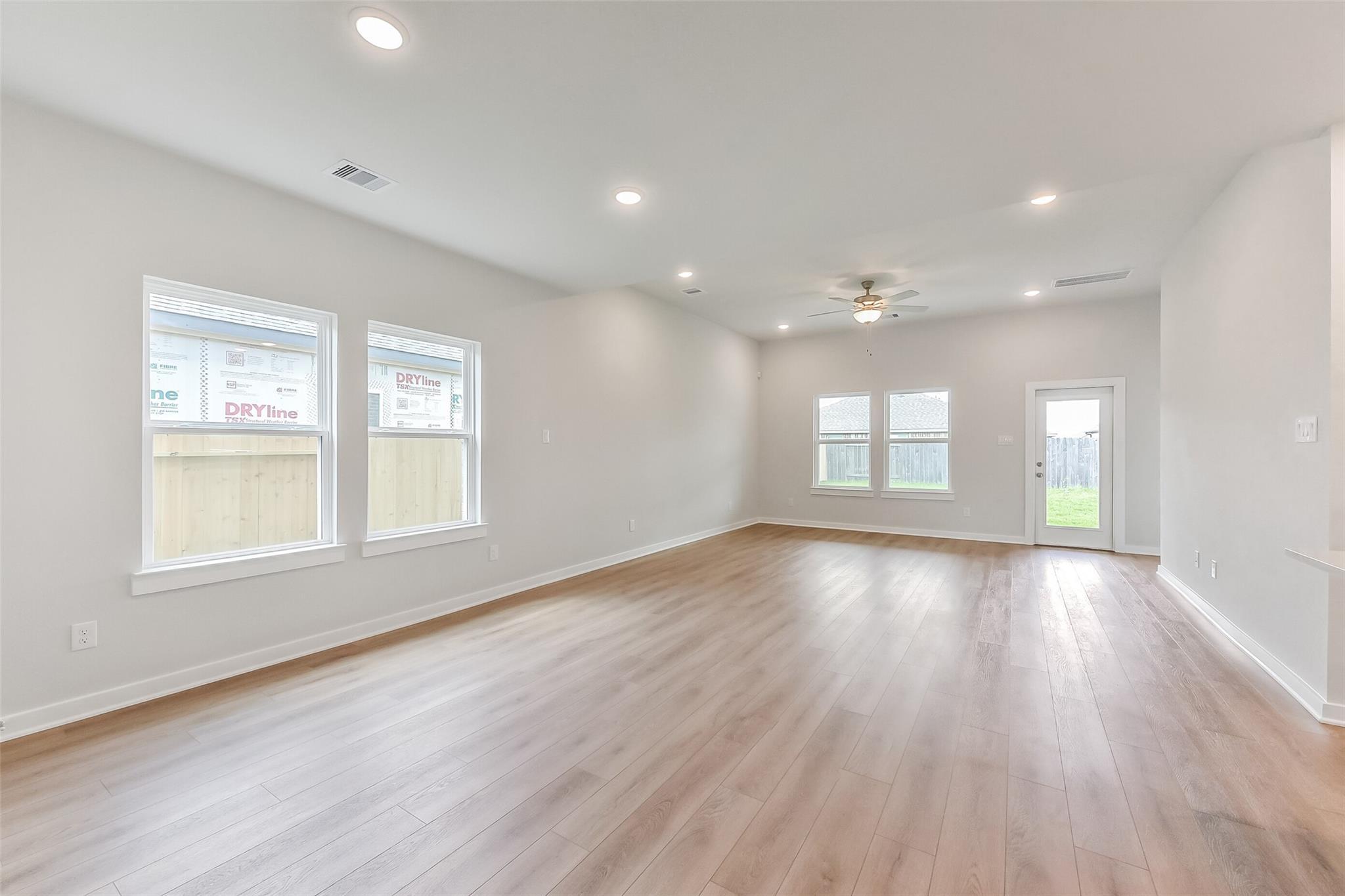 Spacious empty living room with light hardwood floors, large windows, ceiling fan, and glass door to fenced backyard in Davidson Homes The Tierra C, Dayton Texas