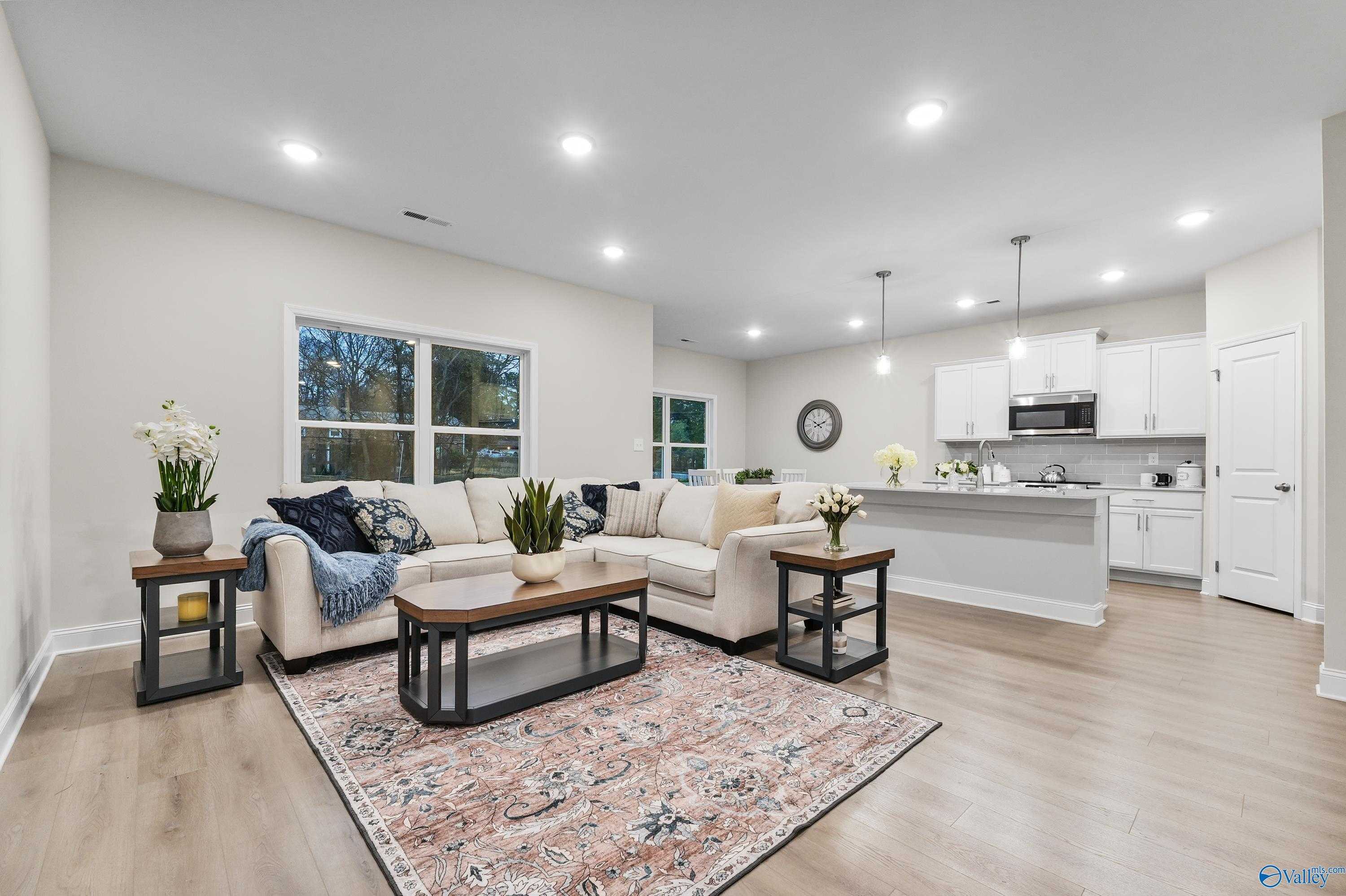 Open-concept living room with beige L-shaped sofa, plants, rug, and white kitchen island in Davidson Homes The Asheville C, Huntsville AL