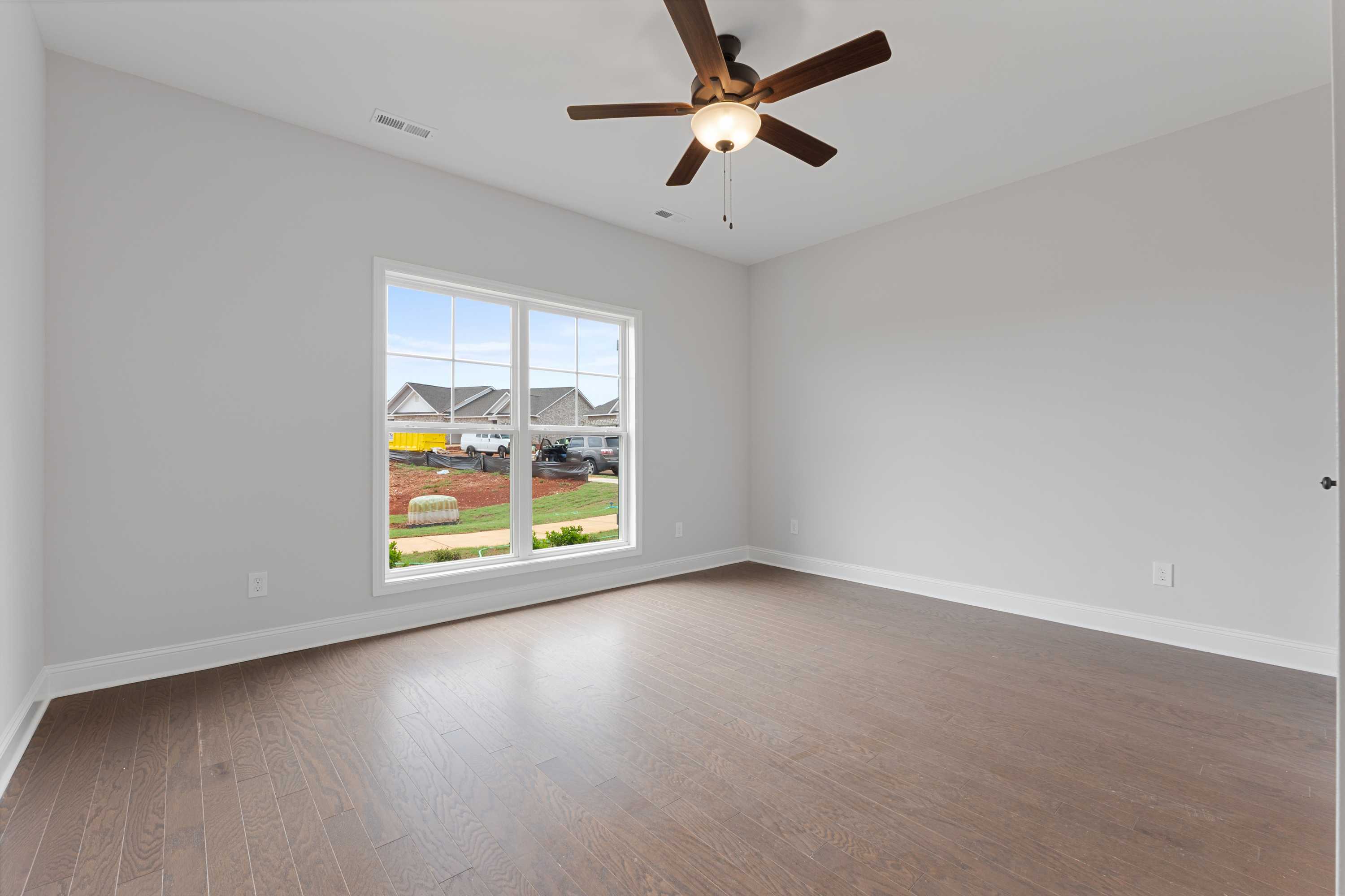 Spacious bedroom in The Copeland D with light gray walls, hardwood floors, ceiling fan, and large windows viewing neighborhood homes