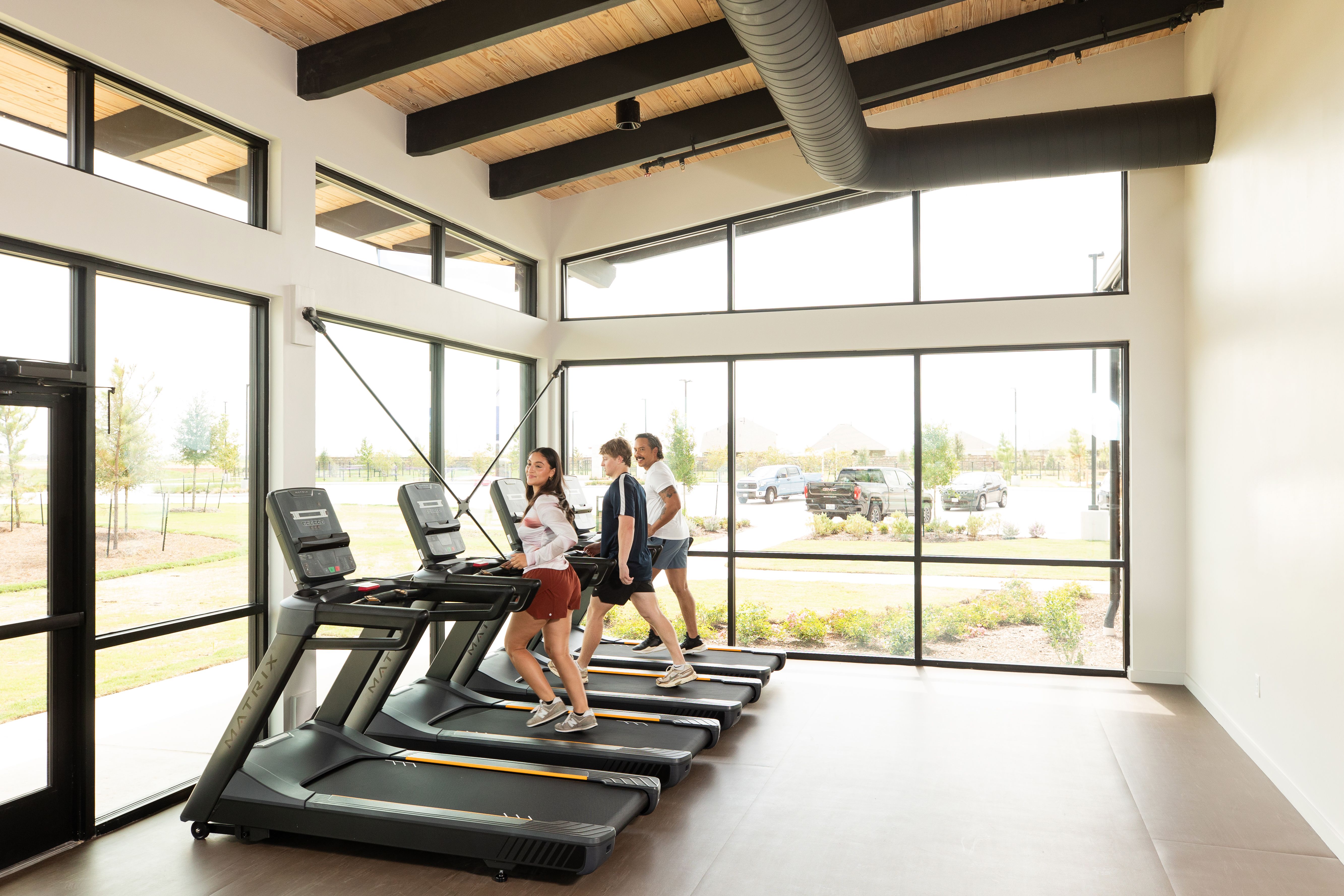 Modern fitness center at Emberly in Beasley, Texas with treadmills, exposed wood beams, and large windows overlooking greenery