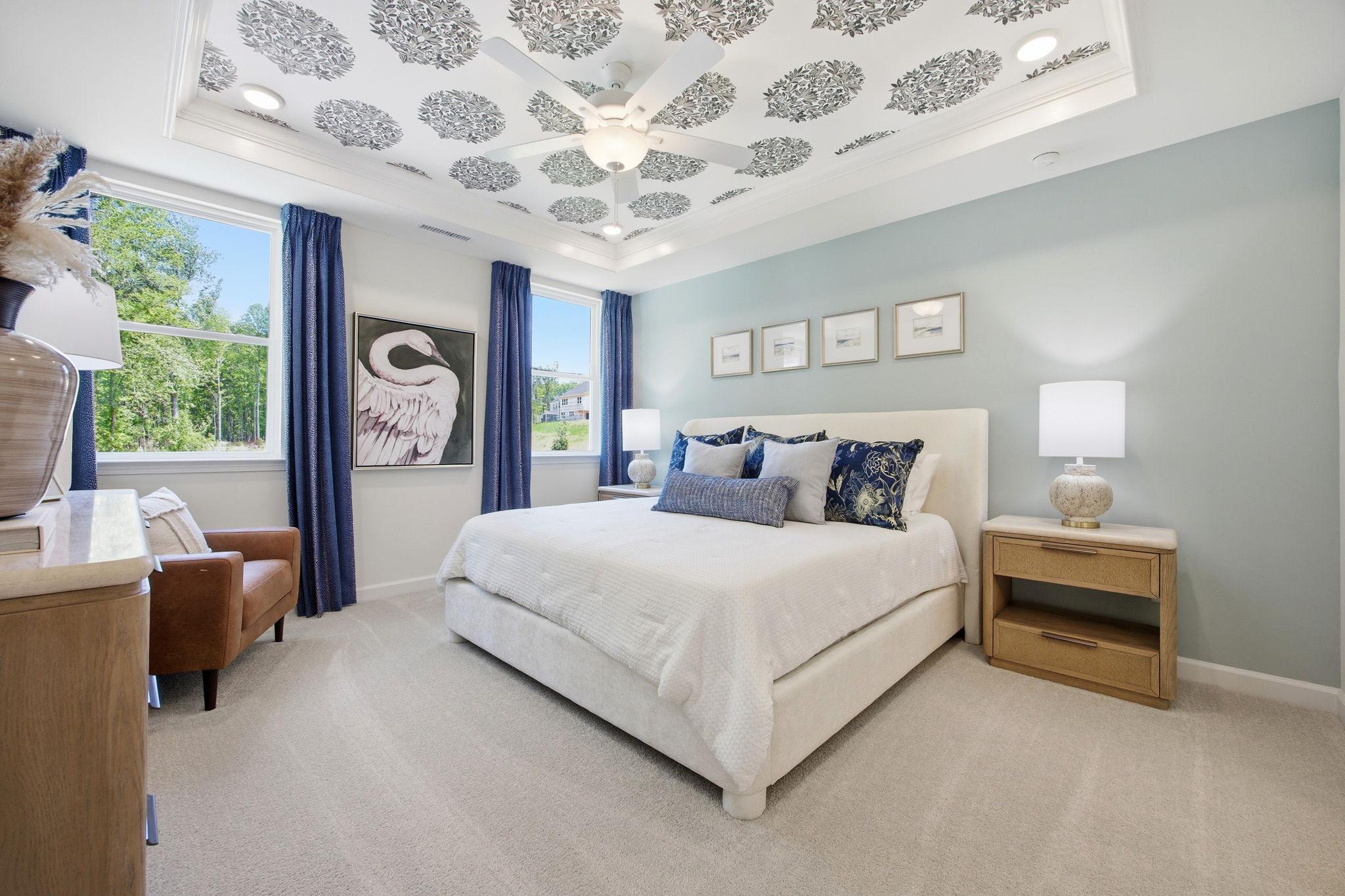 Elegant primary bedroom at Sage on North Main in Wake Forest NC with patterned ceiling, white upholstered bed, blue accents, and serene views