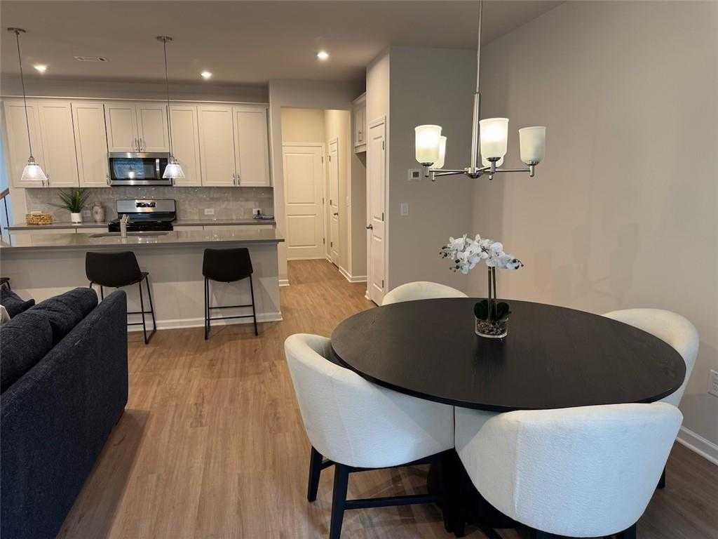 Modern open-concept dining and living space with round black table, white chairs, gray sectional sofa, and white kitchen cabinets in Davidson Homes The Cary A, Kennesaw