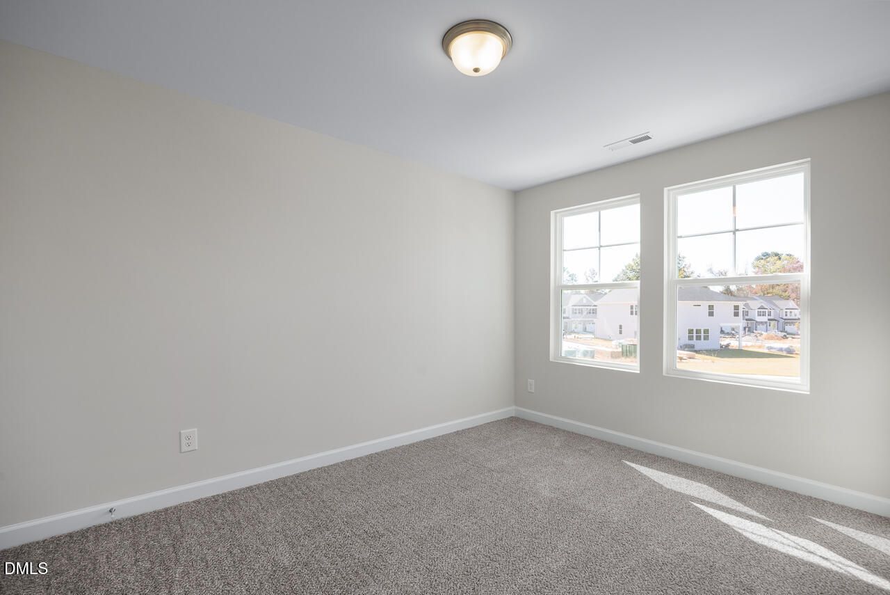 Bright empty bedroom with neutral walls, gray carpet, and dual windows overlooking neighborhood in The Grace C, Lillington, NC