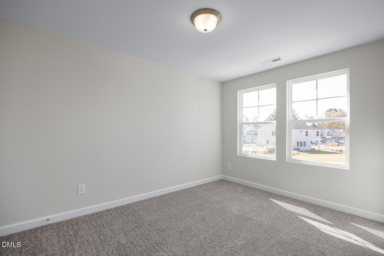 Bright empty bedroom with neutral walls, gray carpet, and dual windows overlooking neighborhood in The Grace C, Lillington, NC