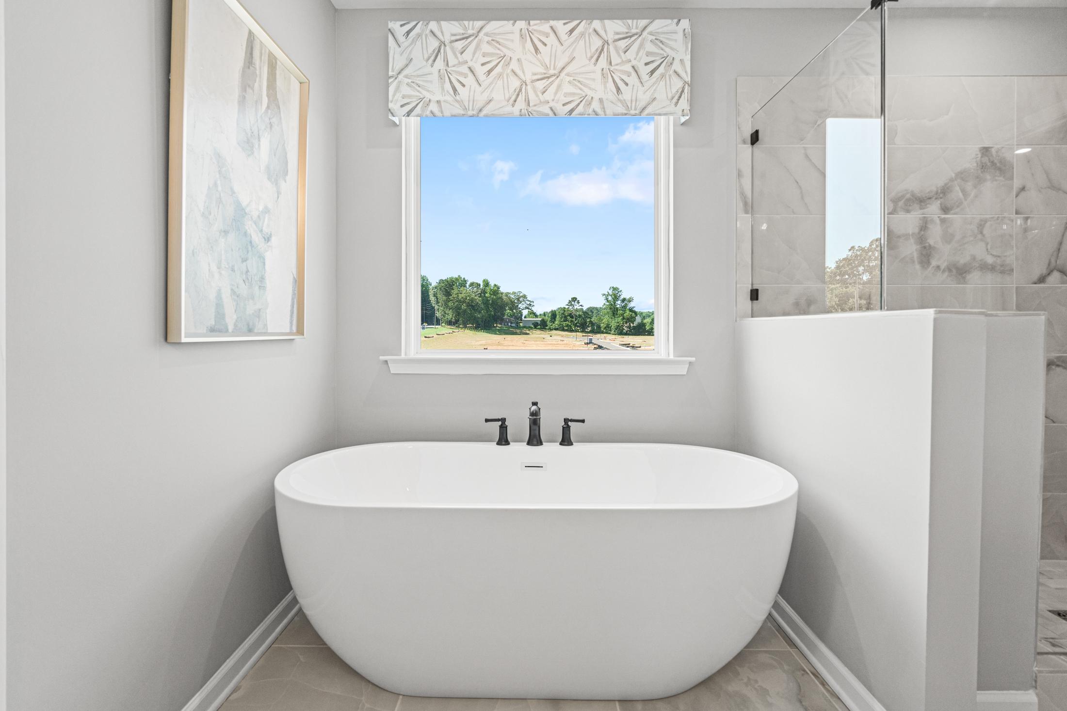 Spacious master bathroom in The Hickory B with freestanding white tub, large window view of trees, and glass shower