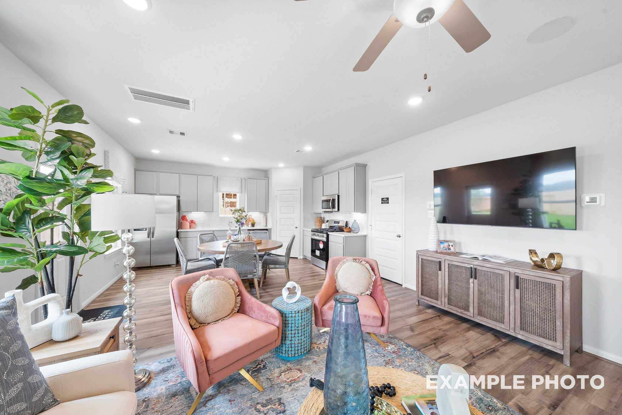 Modern open-concept living room in The Trinity showcasing pink armchairs, blue vase, hardwood floors, white kitchen, and wall TV
