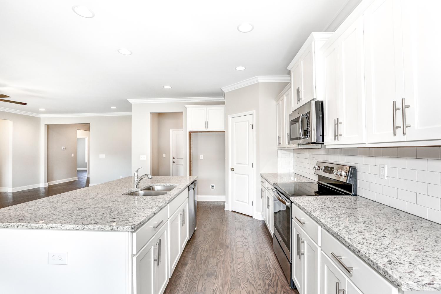 Spacious open kitchen in The Montgomery home featuring white shaker cabinets, granite island with sink, stainless steel appliances, subway tile backsplash