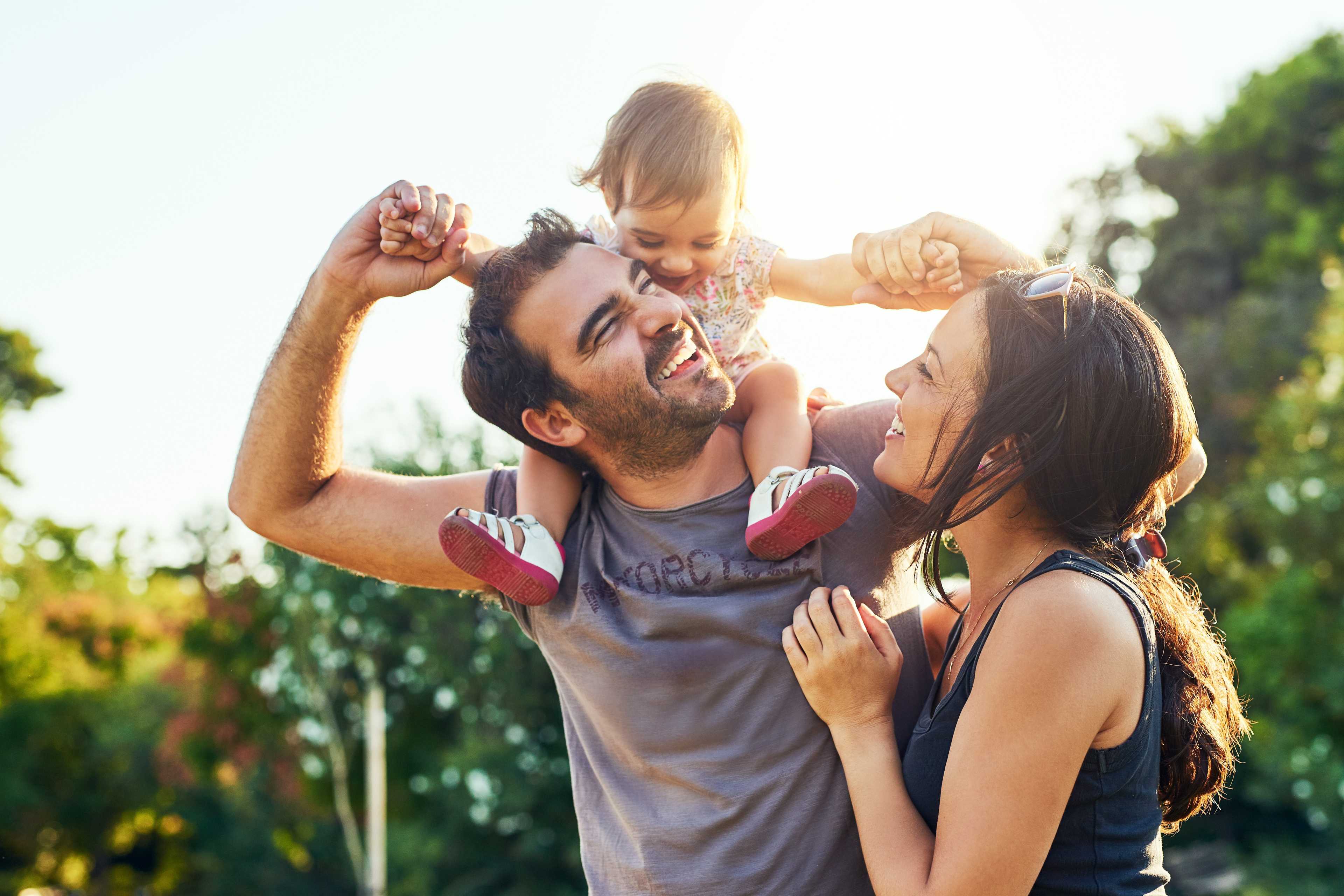 Happy family in sunny park: father with baby on shoulders, smiling mother beside at Spring Branch Crossing, Conroe TX