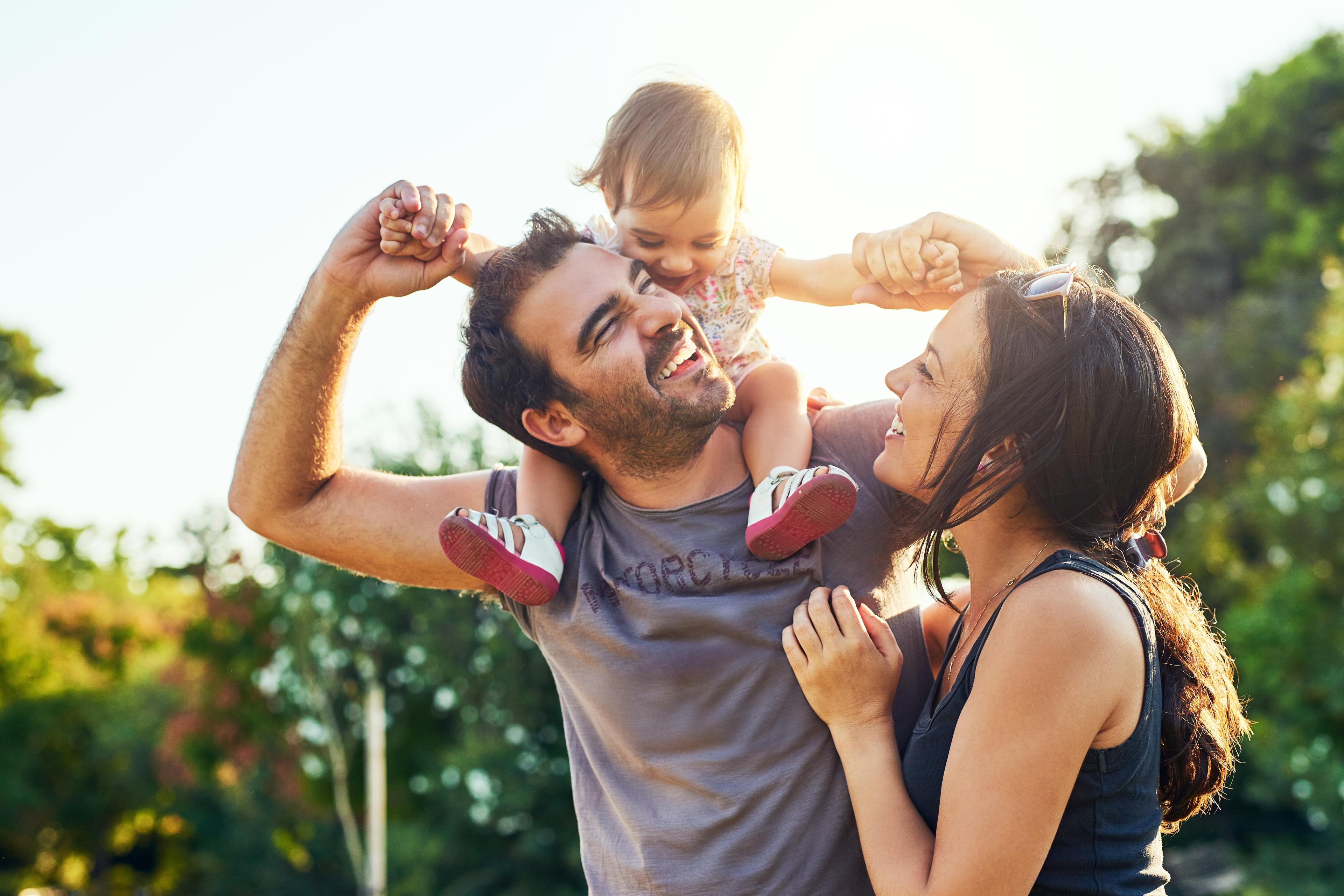 Happy family in sunny park: father with baby on shoulders, smiling mother beside at Spring Branch Crossing, Conroe TX