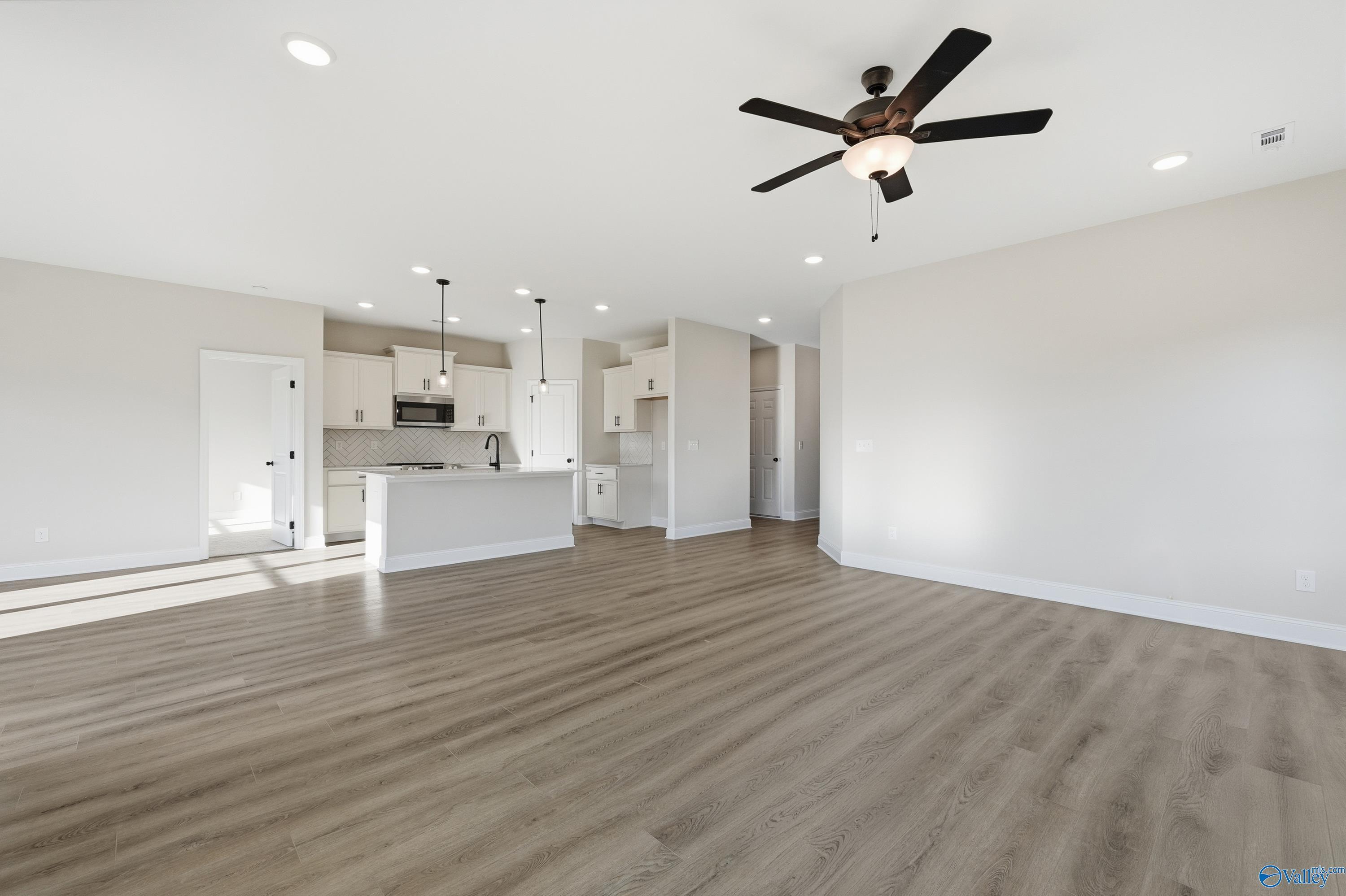 Open-concept kitchen with white cabinets, large island sink, and adjacent living area featuring light wood floors and ceiling fans in The Franklin E, Hazel Green