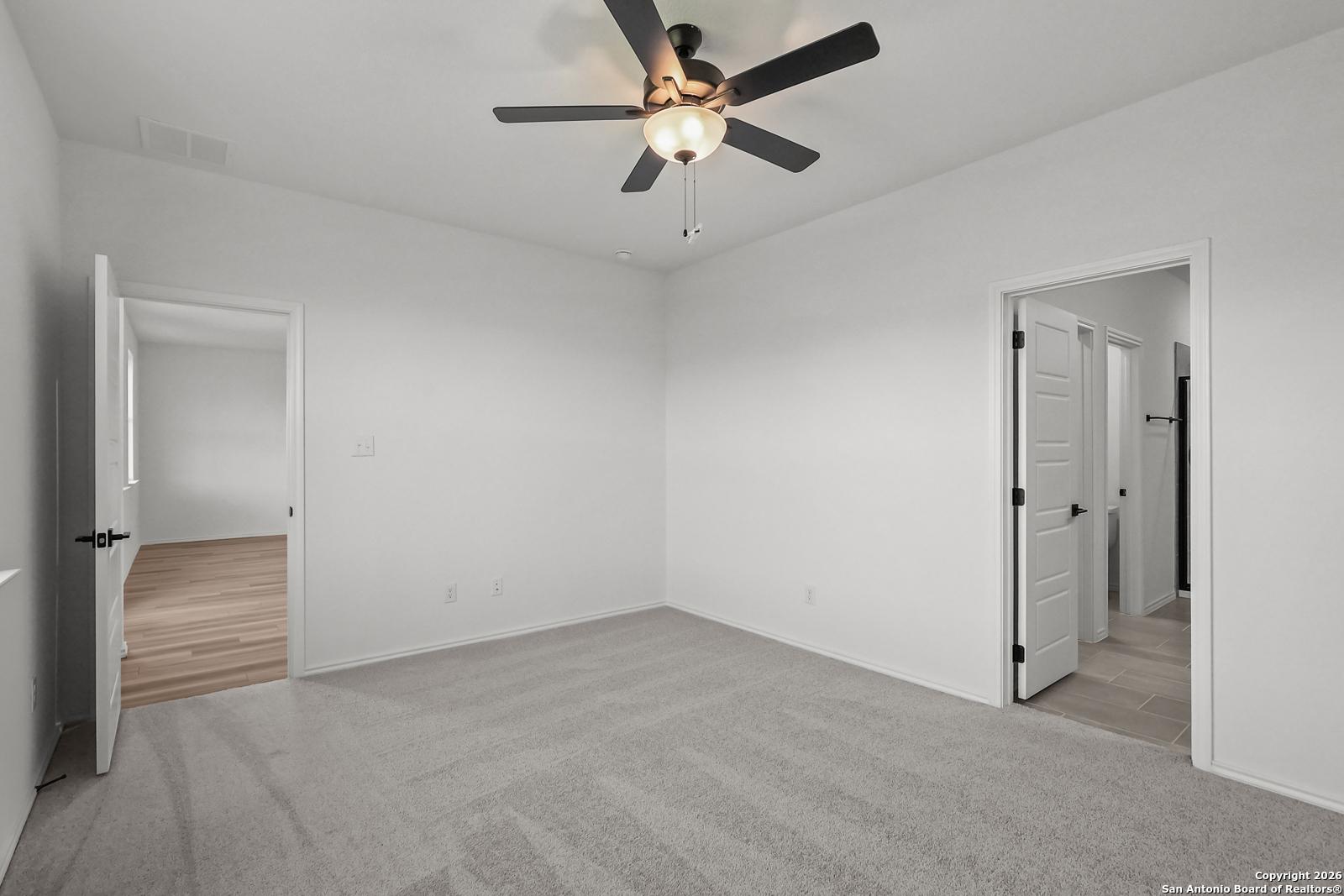 Bright bedroom with ceiling fan, white walls, gray carpet, and open doors to adjacent rooms in Davidson Homes The Douglas E, San Antonio