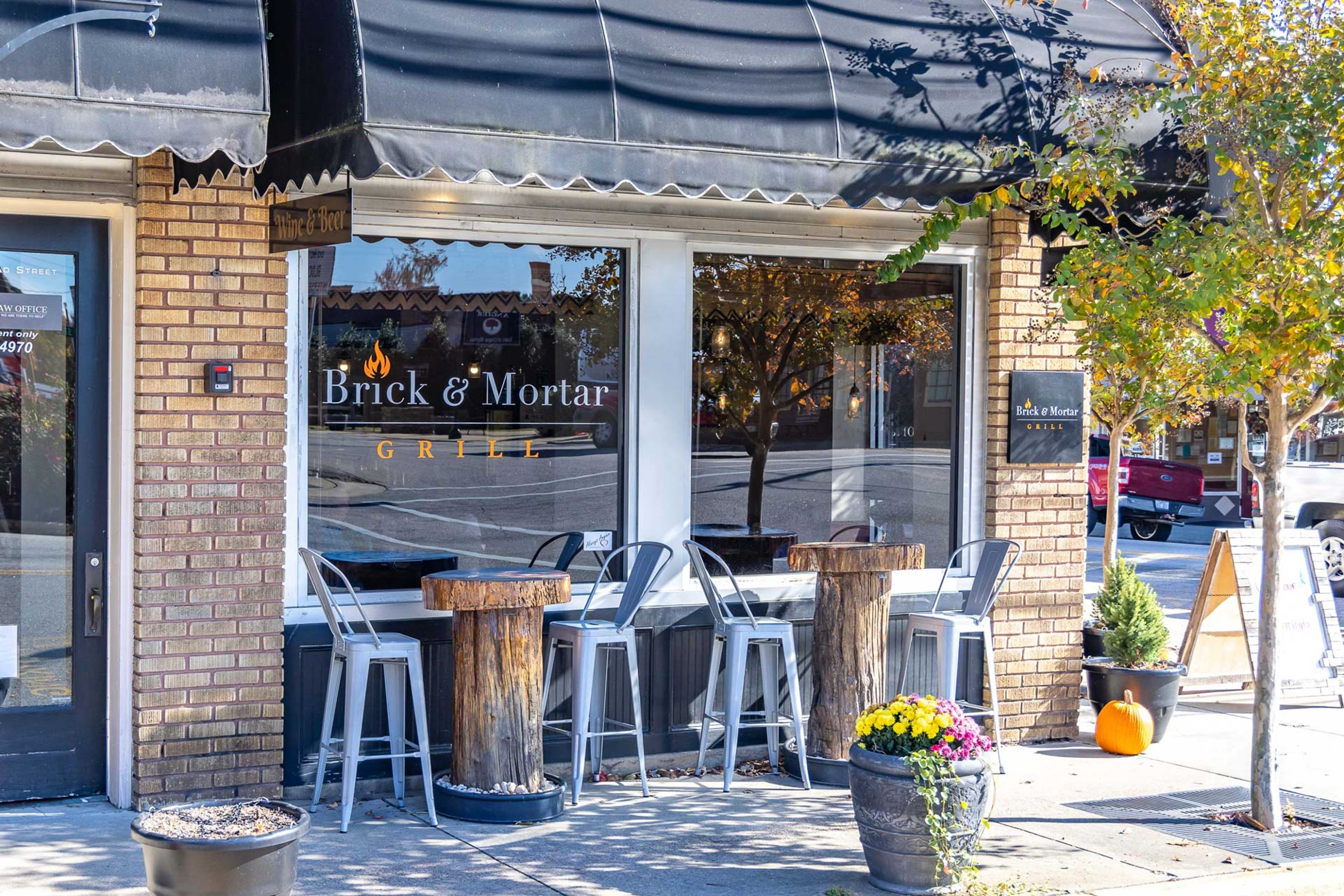 Charming Brick & Mortar Cafe exterior in Angier NC with brick facade, black awning, outdoor barrel tables, metal stools, fall flowers and pumpkins near Weatherford East