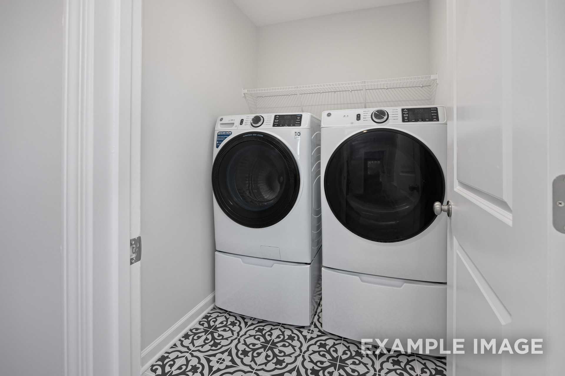 Modern laundry room in The Grace C with white front-load washer and dryer, overhead shelf, and patterned tile floor