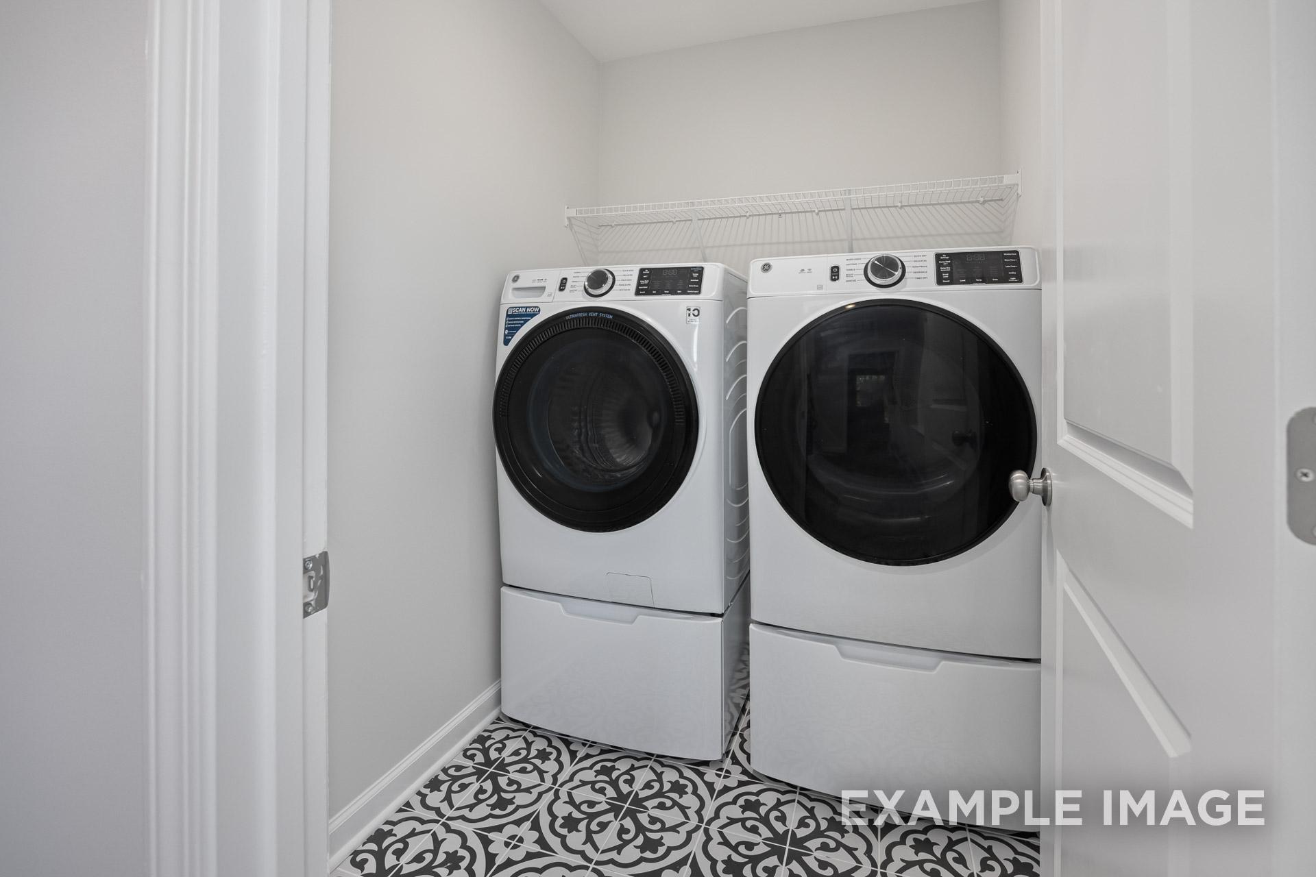 Modern laundry room in The Grace C with white front-load washer and dryer, overhead shelf, and patterned tile floor
