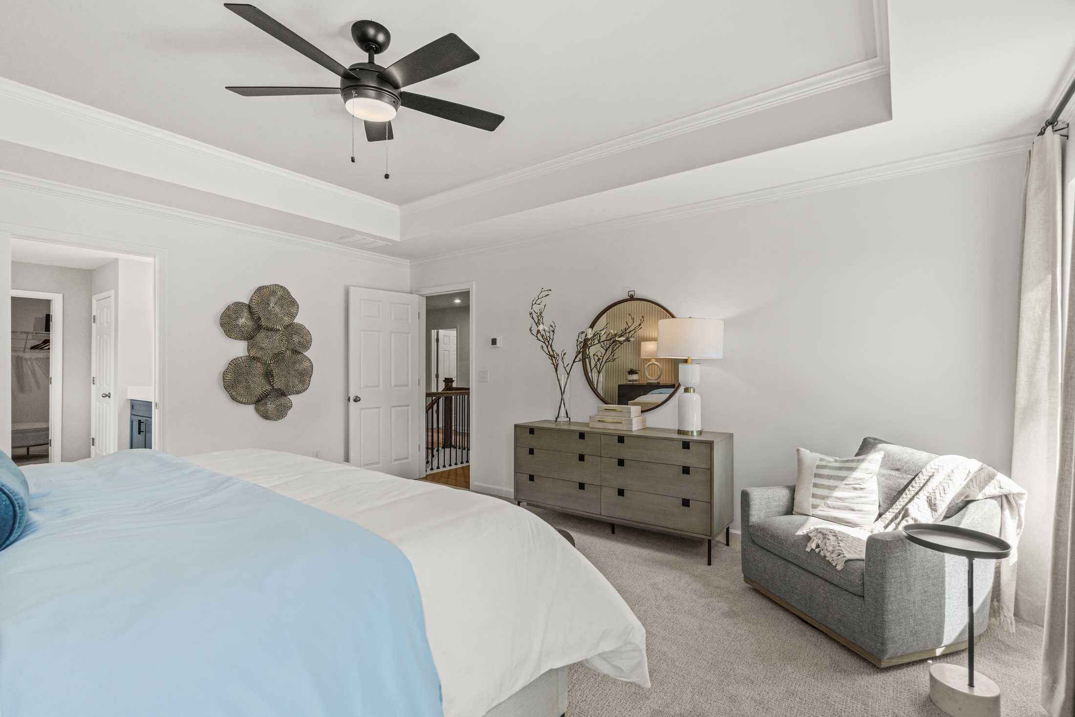 Spacious master bedroom in The Hickory A with king bed, gray dresser, armchair, ceiling fan, and round mirror
