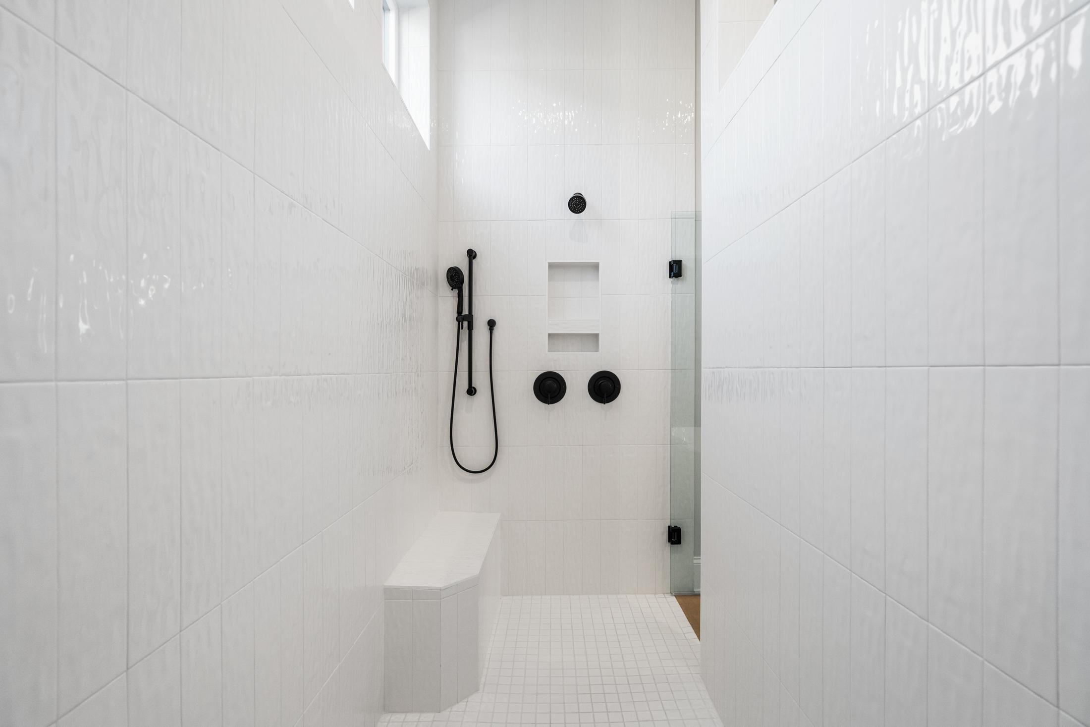 Modern white subway tile walk-in shower with black rain showerhead, handheld sprayer, bench, and niche in The Seaside master bath