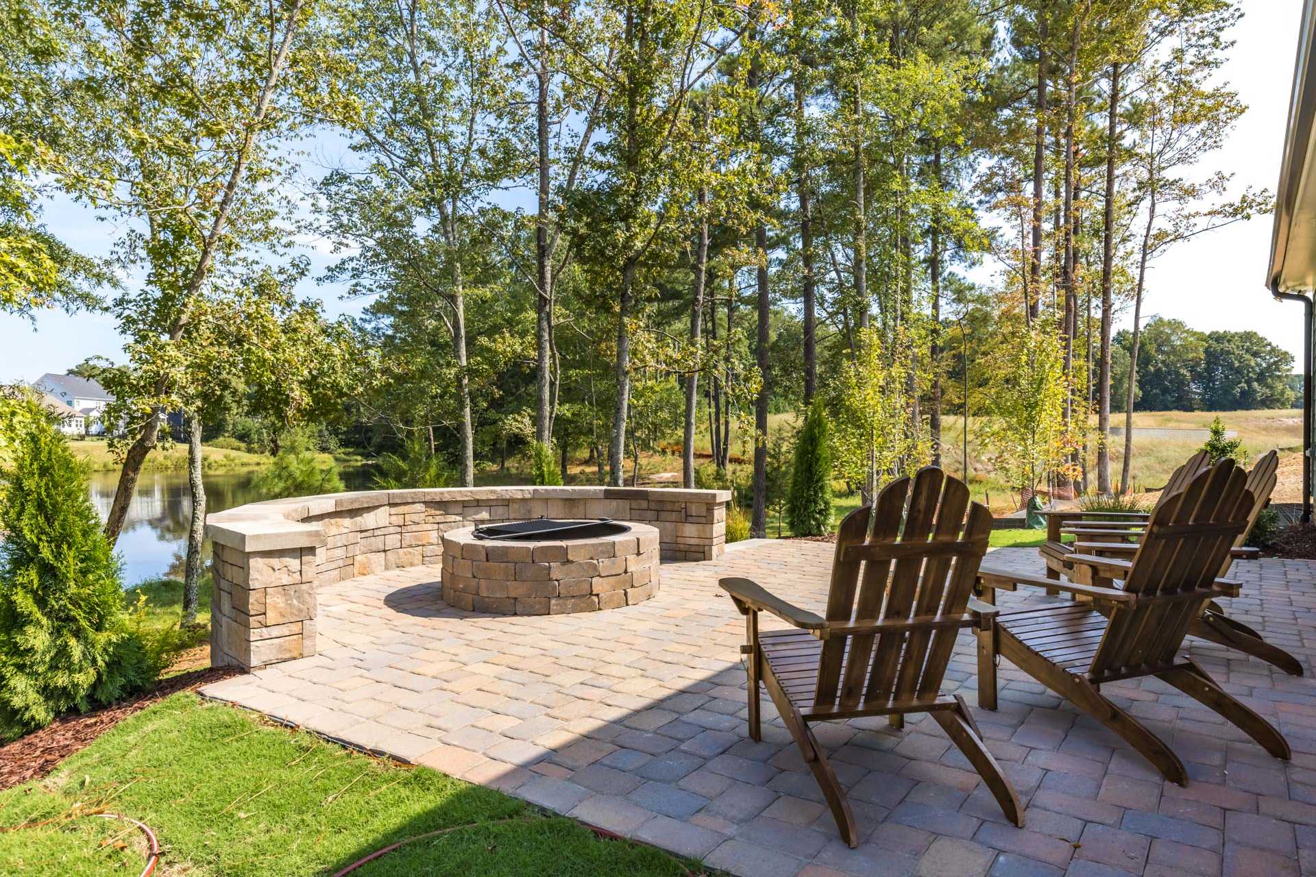 Cozy stone fire pit on paver patio at Glenmere in Knightdale NC with Adirondack chairs, trees, and pond view
