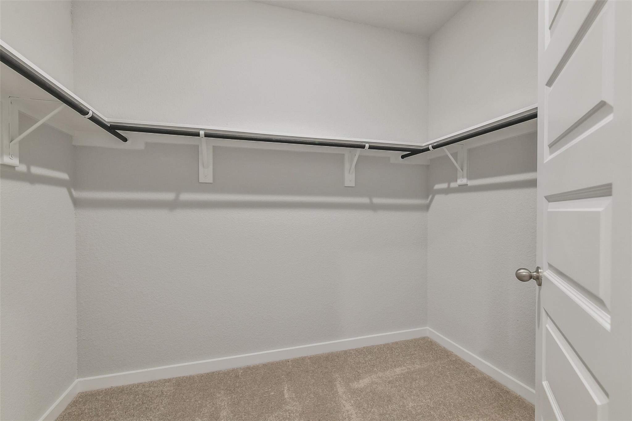 Spacious walk-in closet with white walls, beige carpet, and wooden hanging rods in Davidson Homes The Brazos E, Cleveland, Texas