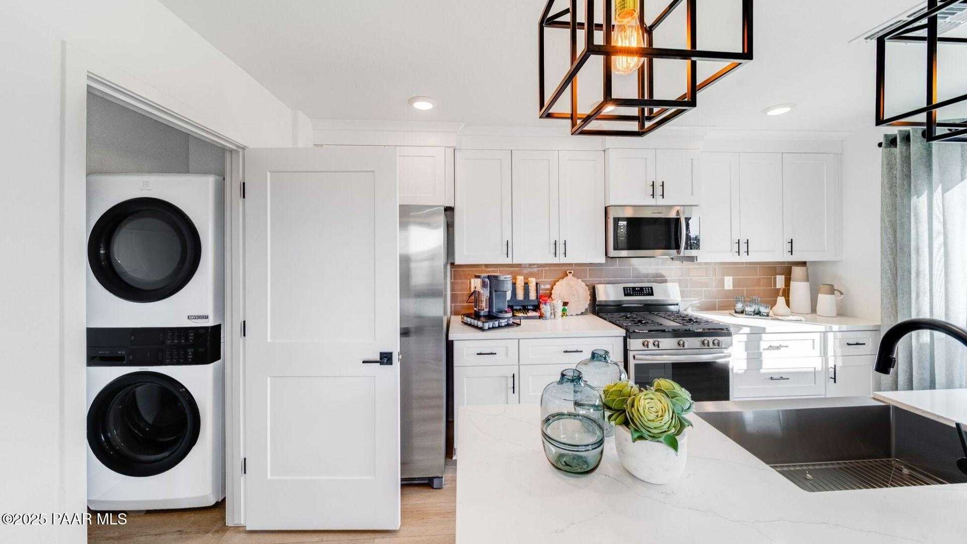 Modern kitchen with white cabinets, stainless steel appliances, and adjacent laundry room featuring stacked washer dryer in The Wilmington B, Prescott AZ