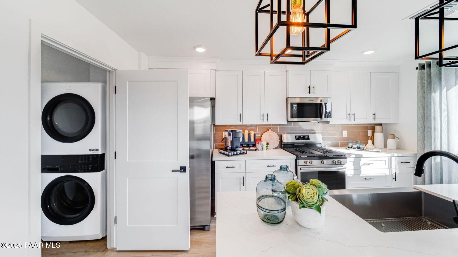 Modern kitchen with white cabinets, stainless steel appliances, and adjacent laundry room featuring stacked washer dryer in The Wilmington B, Prescott AZ