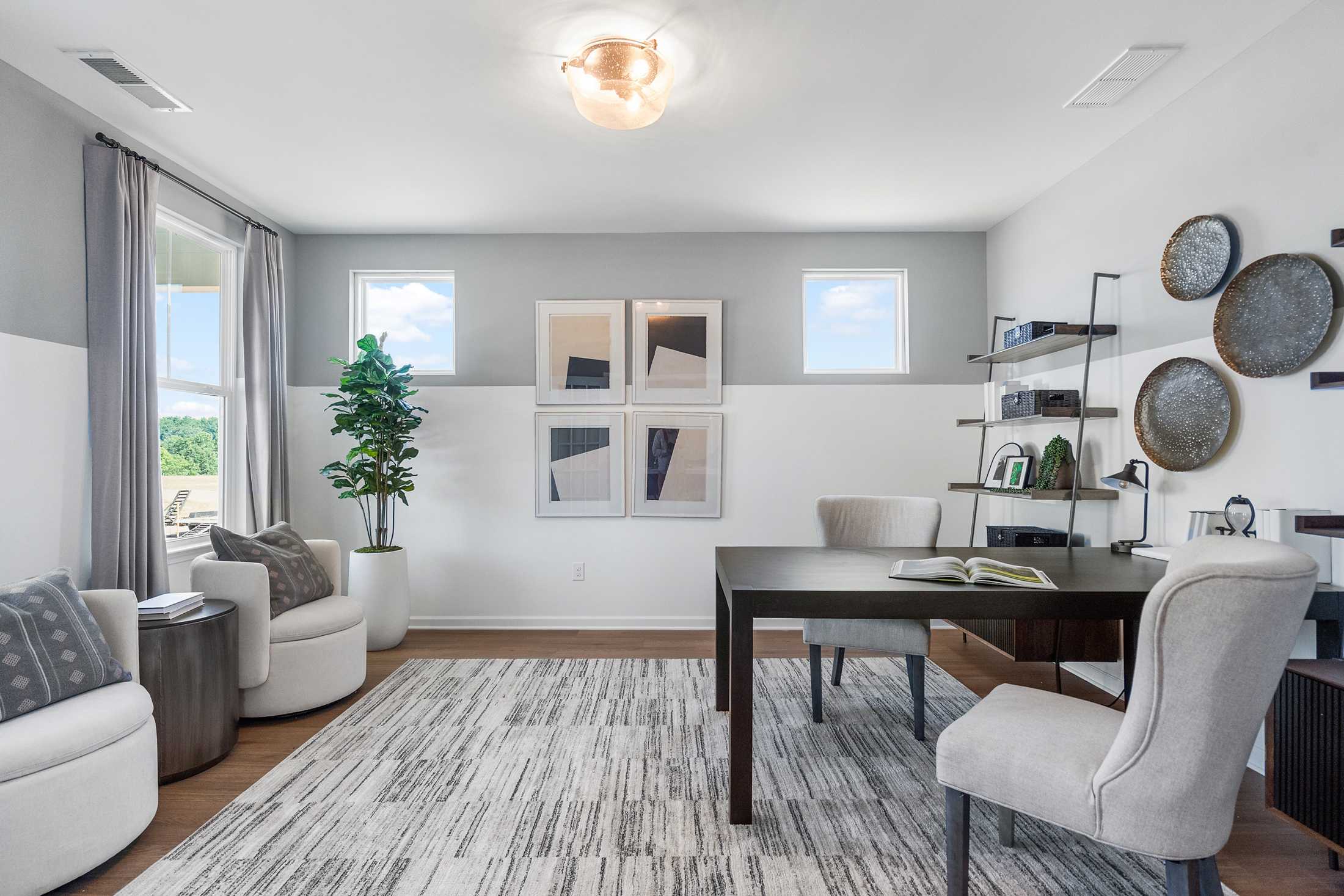 Spacious home office in The Hickory A featuring wooden desk, white armchairs, bookshelves, abstract art, and large windows