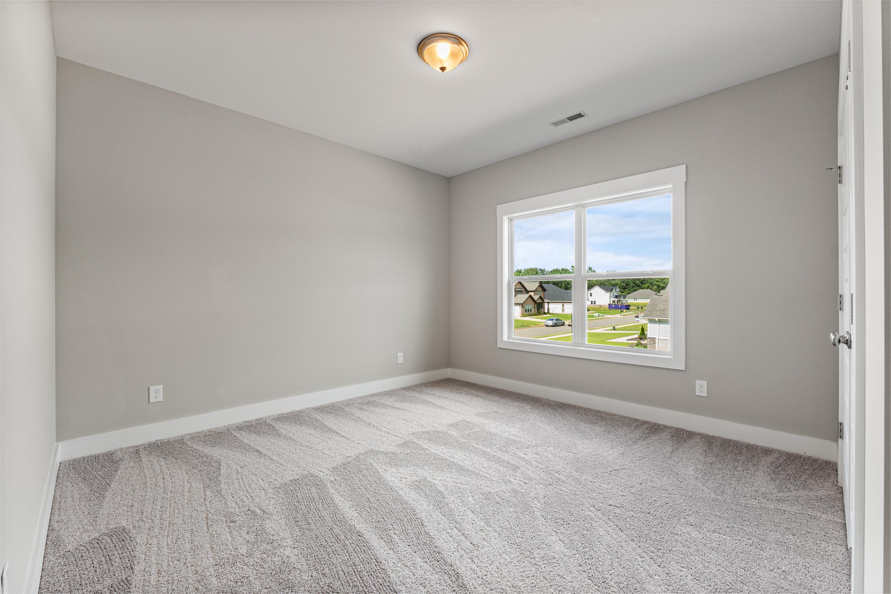 Spacious empty bedroom in The Oxford C home with light gray walls, plush carpet, and window view of suburban neighborhood