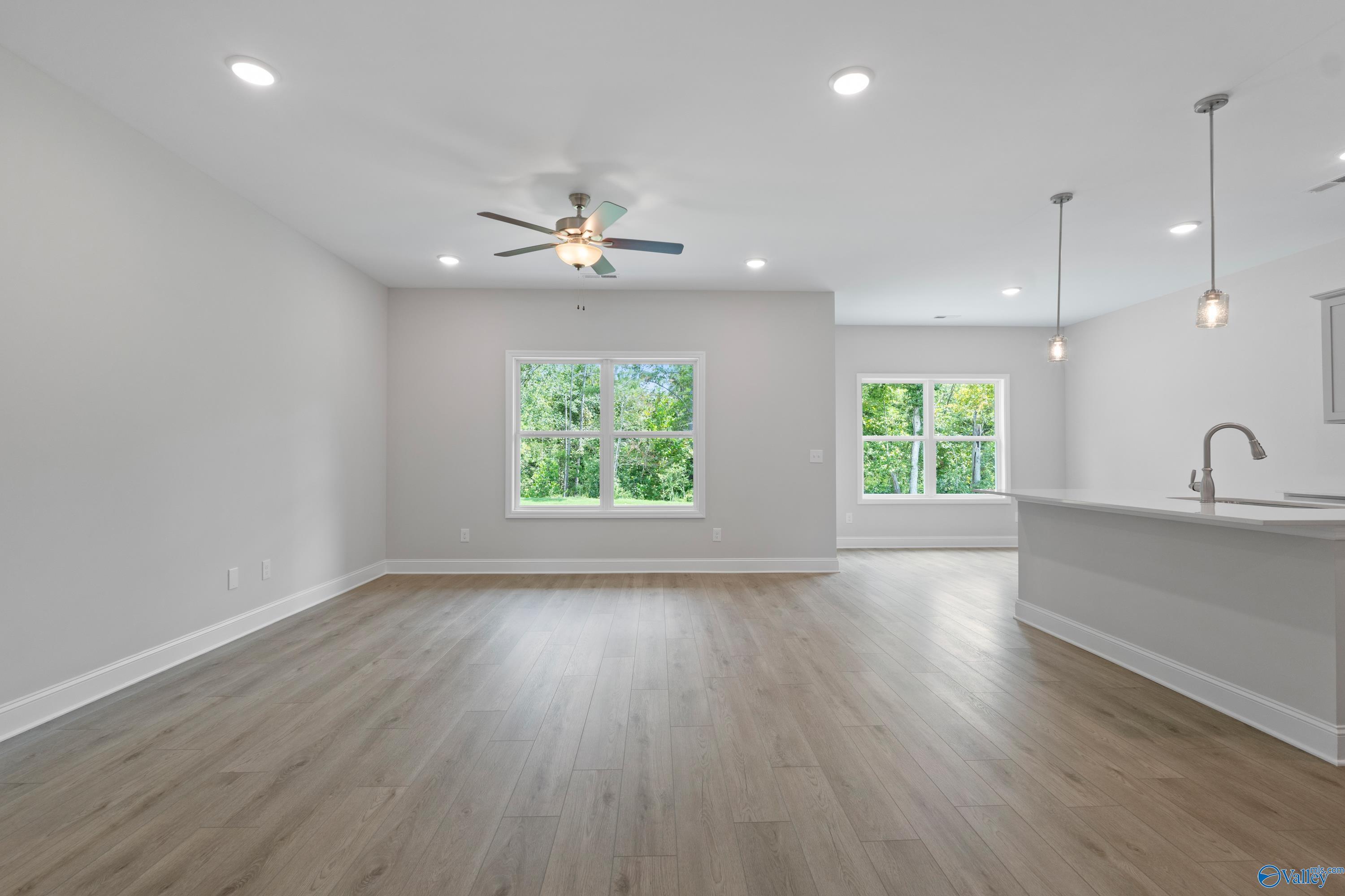 Open-concept living room with hardwood floors, ceiling fan, and large windows overlooking trees in The Asheville by Davidson Homes, Huntsville AL
