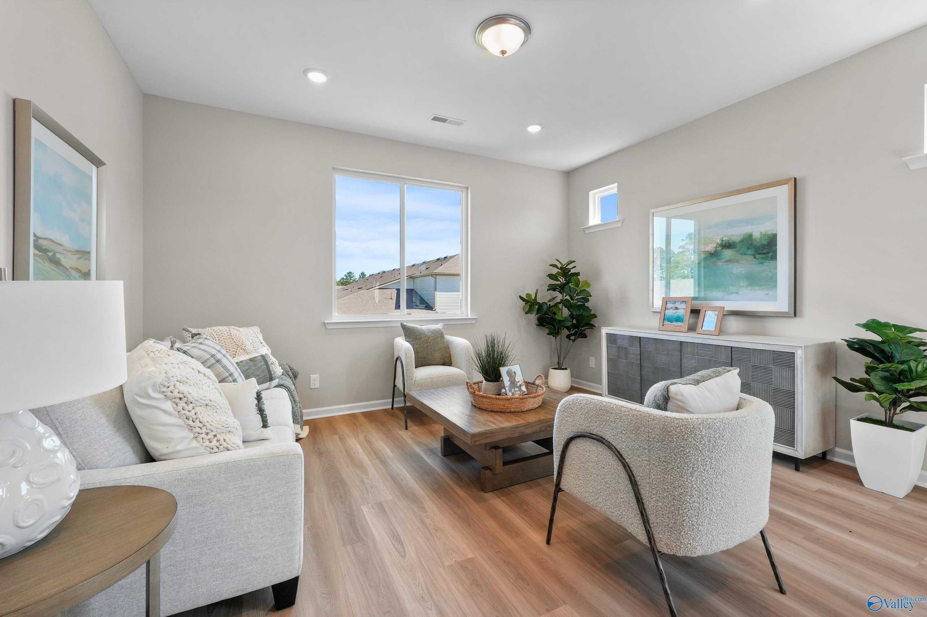 Cozy living room featuring gray sofa, white armchairs, potted plants, and hardwood floors in Evermore Homes The Malibu, Madison Alabama