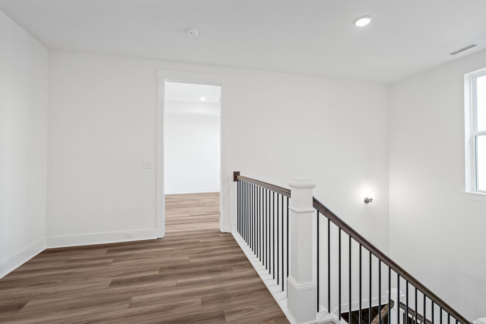 Bright upstairs hallway with hardwood floors, white walls, open doorway and balcony railing in The Alston A home, Murfreesboro, TN