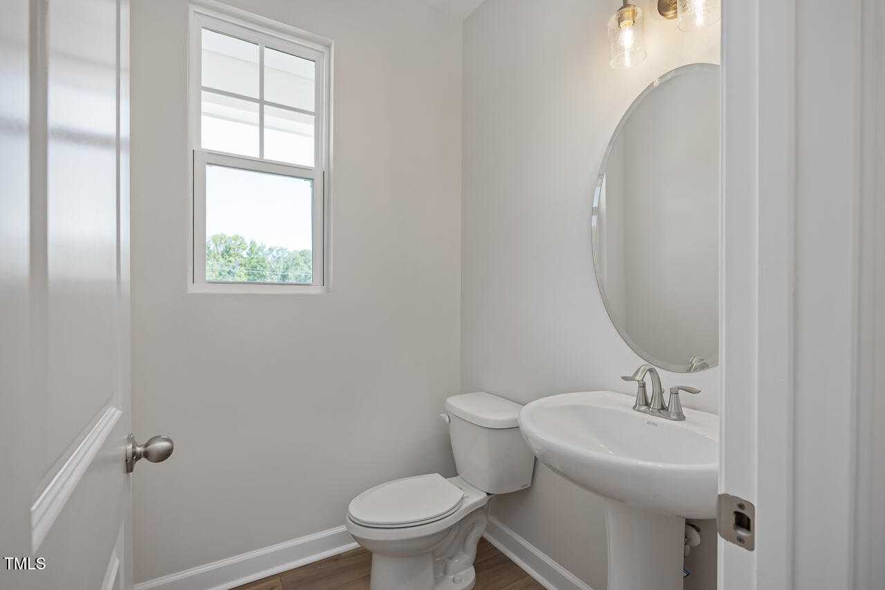 Modern half bath with round mirror, white pedestal sink, toilet, and window in Davidson Homes The Graham Interior, Fuquay-Varina, NC