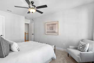 Spacious master bedroom in The Washington H townhome by Davidson Homes, white bedding, gray accent chair, ceiling fan, abstract wall art