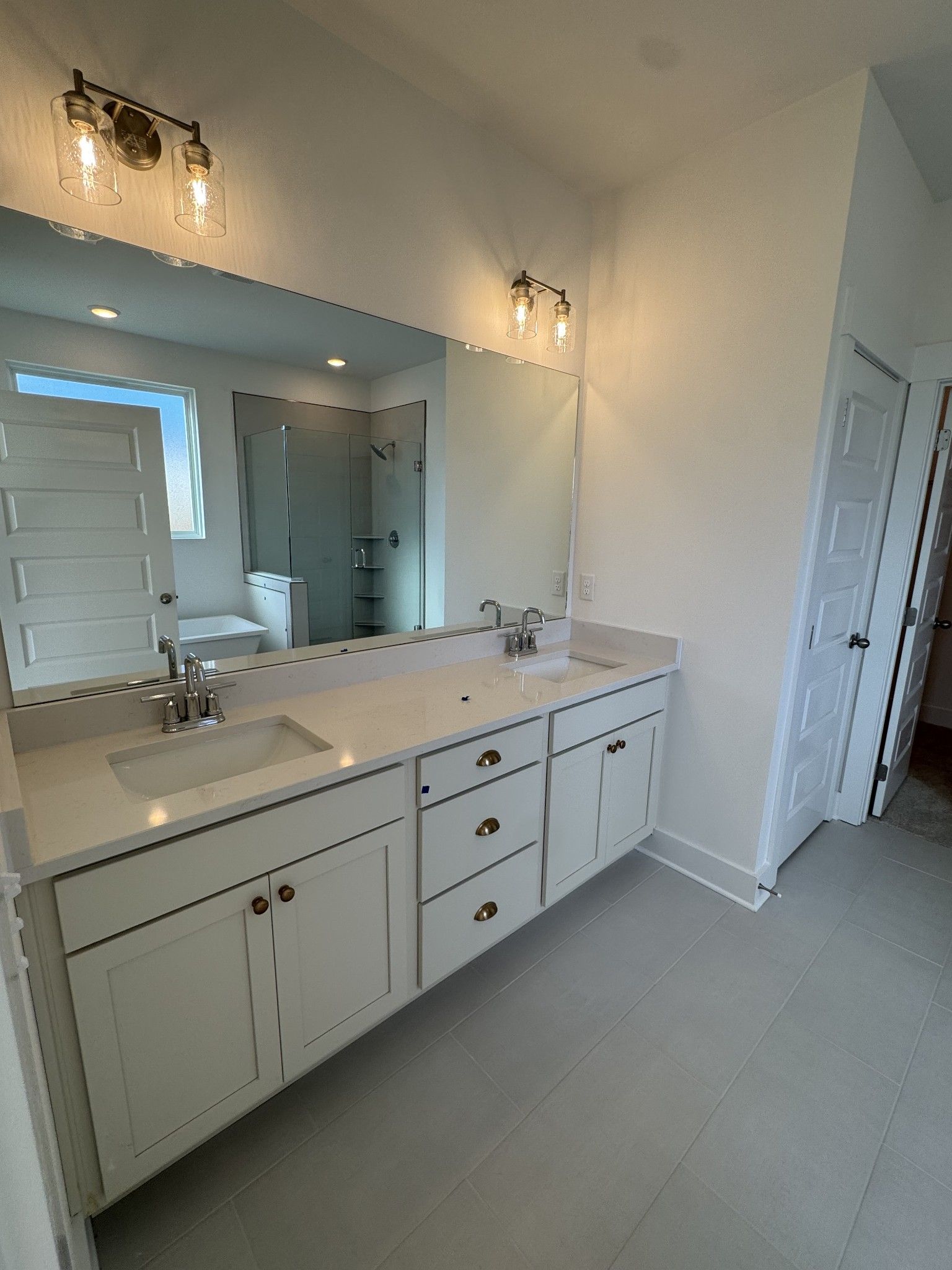 Modern primary bathroom with double vanity, white shaker cabinets, quartz counters, large mirror, and glass shower in Davidson Homes The Ridgeport, Gallatin, TN