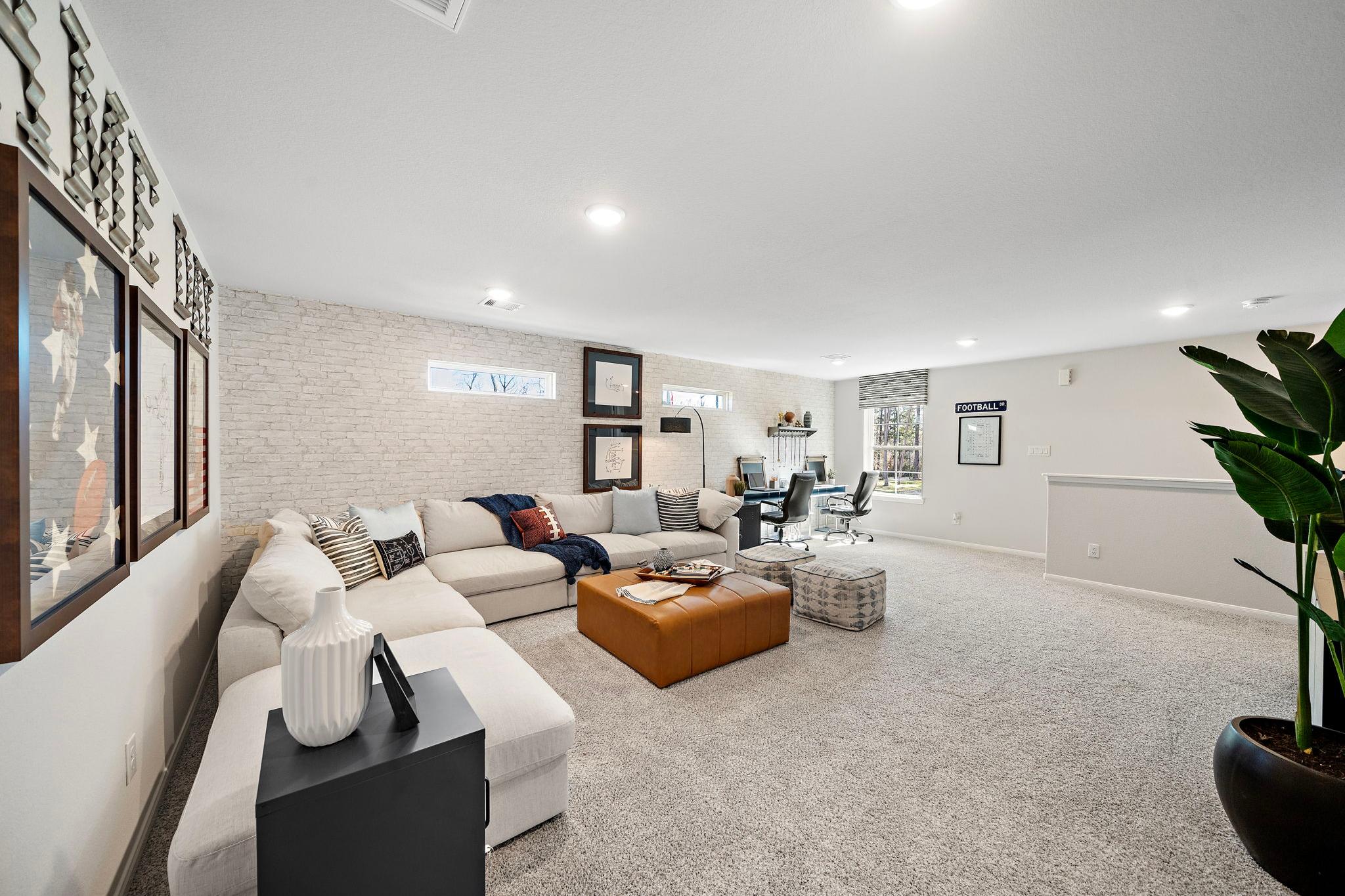 Spacious modern family room at Lakes at Black Oak in Magnolia Texas with L-shaped sofa, leather ottoman, desk nook, and potted plant