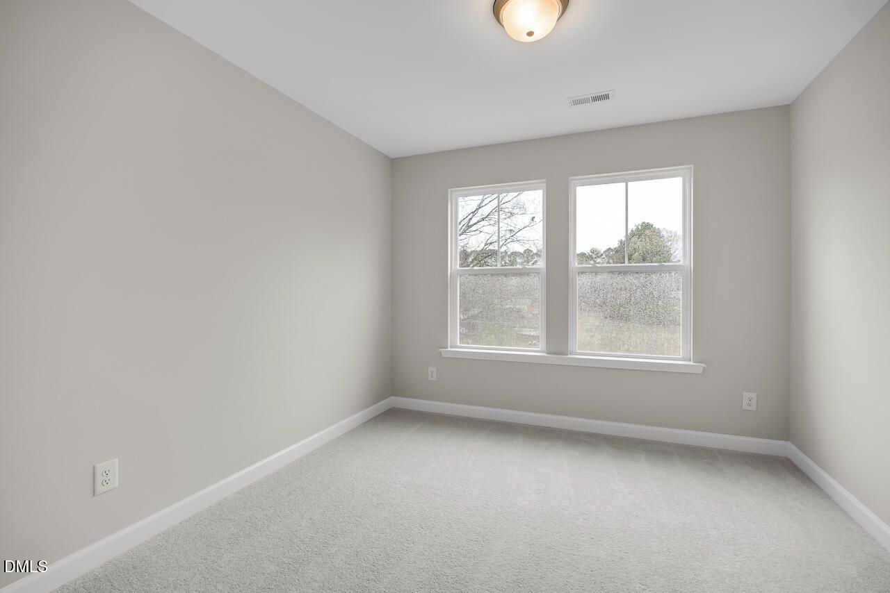 Empty bedroom with light gray walls, large double windows overlooking trees, gray carpet, and ceiling light in Davidson Homes The Avery, Knightdale, NC
