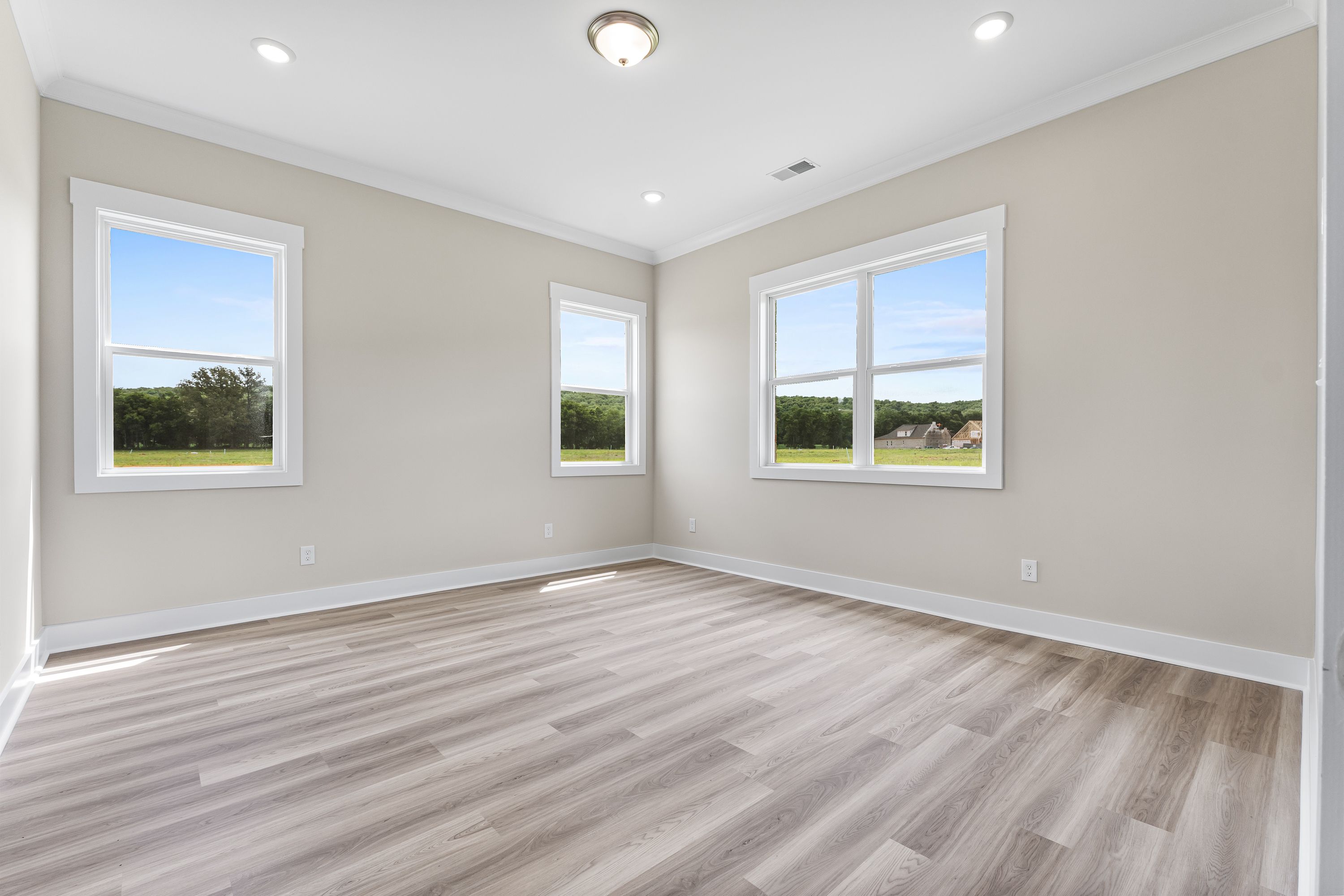 Spacious empty bedroom in The Oxford A with beige walls, light hardwood floors, and large windows overlooking green fields
