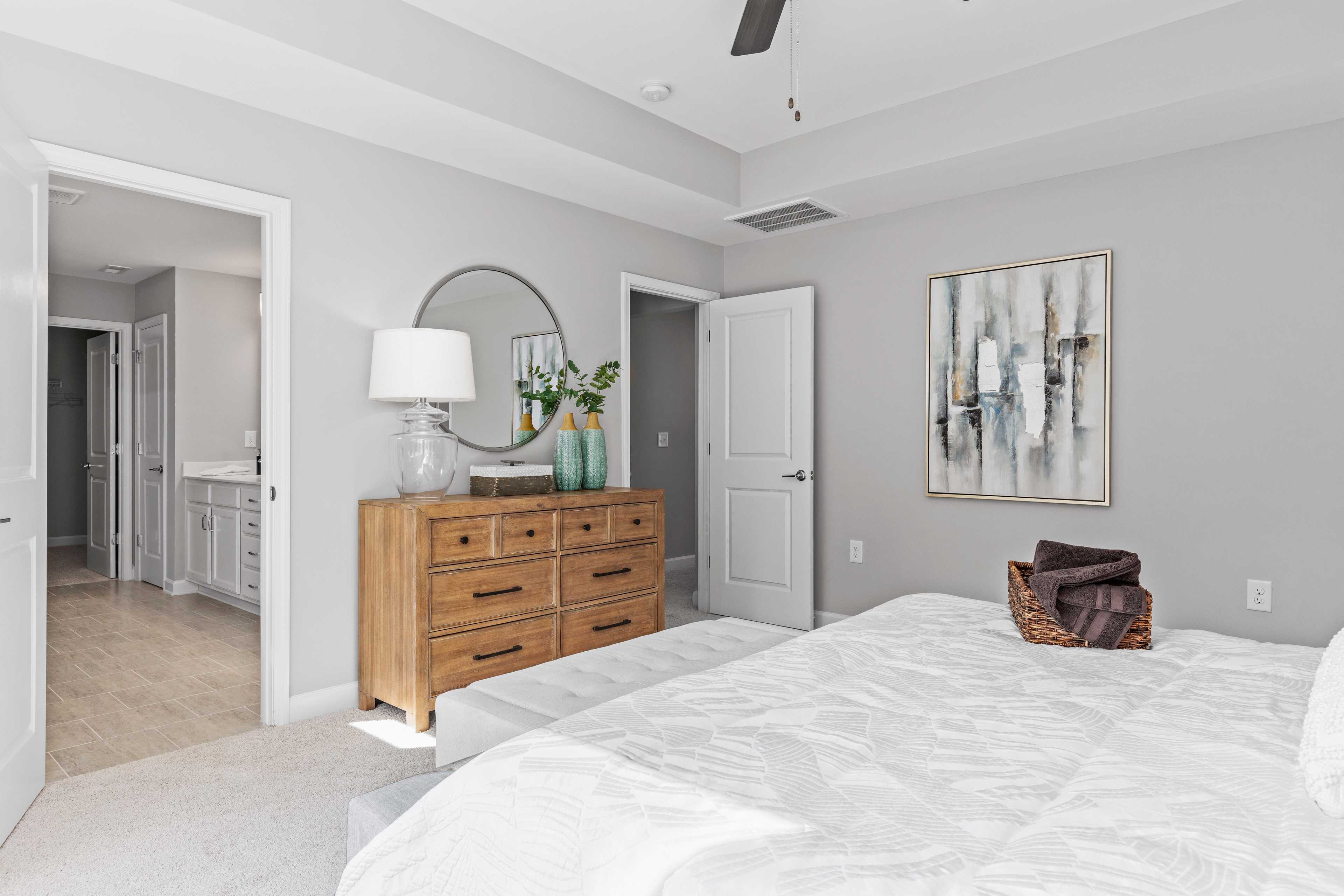 Spacious primary bedroom at Enclave at Belmont in Belmont NC with gray walls, wooden dresser, round mirror, and en-suite bath