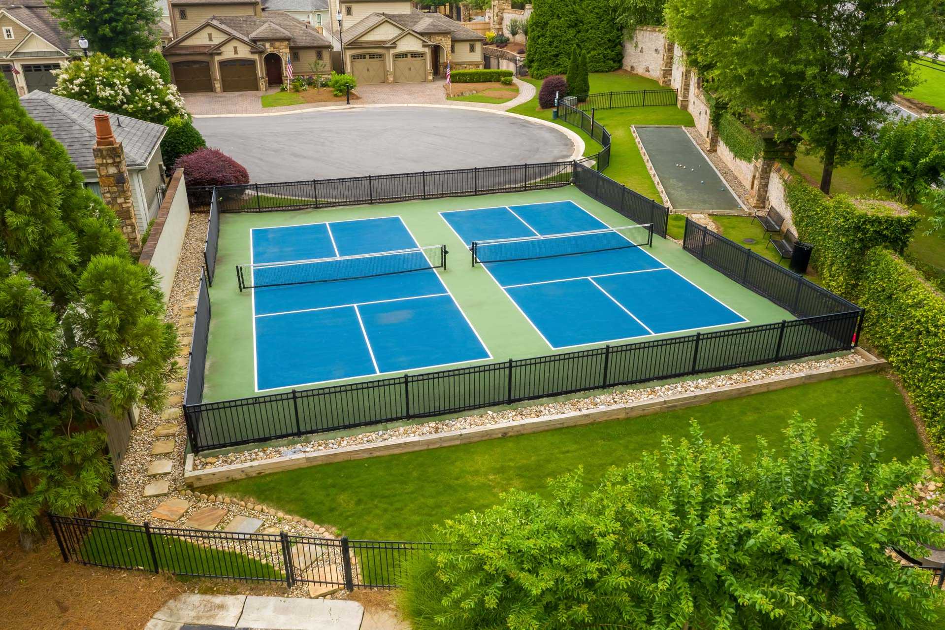 Two blue tennis courts with black fencing at The Village at Towne Lake in Woodstock Georgia amid lush greenery and nearby homes