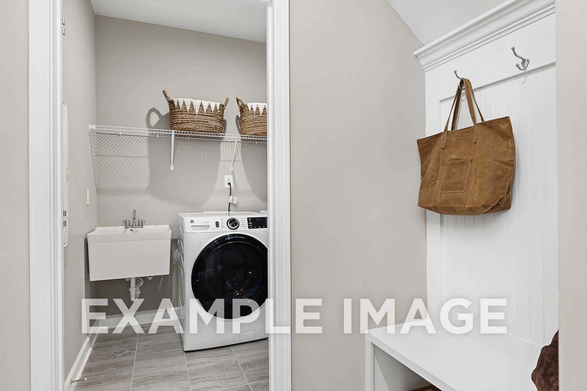 The Ash E laundry room featuring white washer dryer, utility sink, wire shelves with wicker baskets, and coat hooks