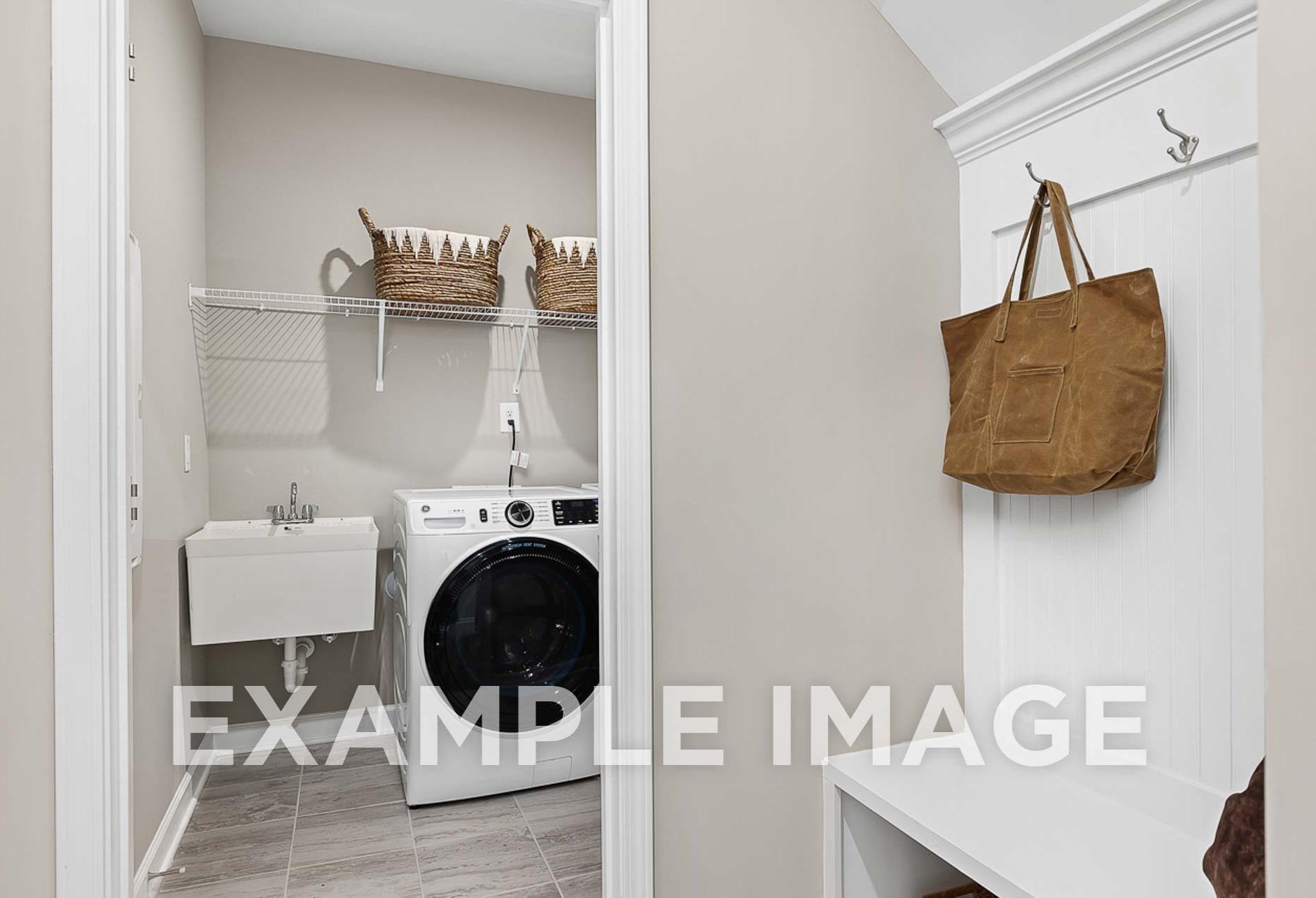 Spacious laundry room in The Ash D featuring white washer-dryer set, utility sink, woven basket shelves, and coat hooks with tote bag