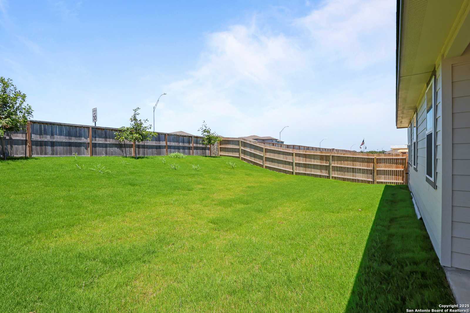 Private fenced backyard with lush green lawn and young trees beside single-story home in Comanche Ridge, San Antonio