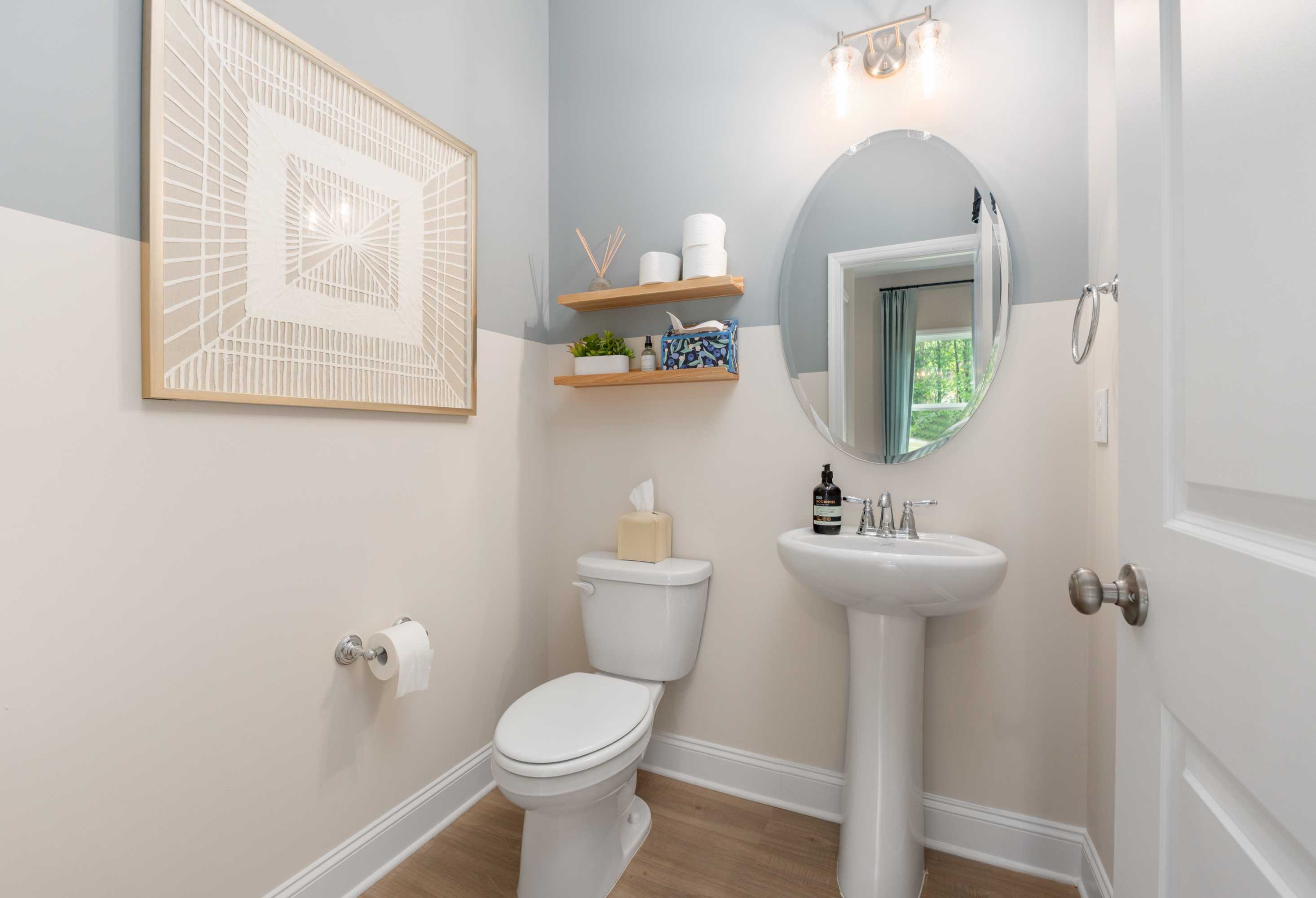 Cozy powder room in The Camden featuring pedestal sink, round mirror, light blue walls, and decorative shelf