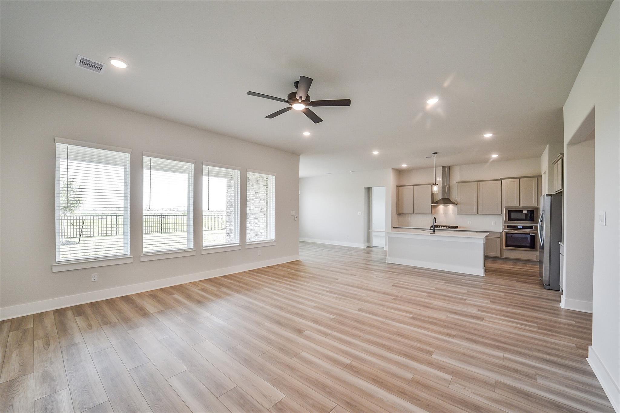 Open-concept living room and kitchen with large windows, white cabinets, island, and ceiling fan in Davidson Homes The George B, Rosharon, Texas