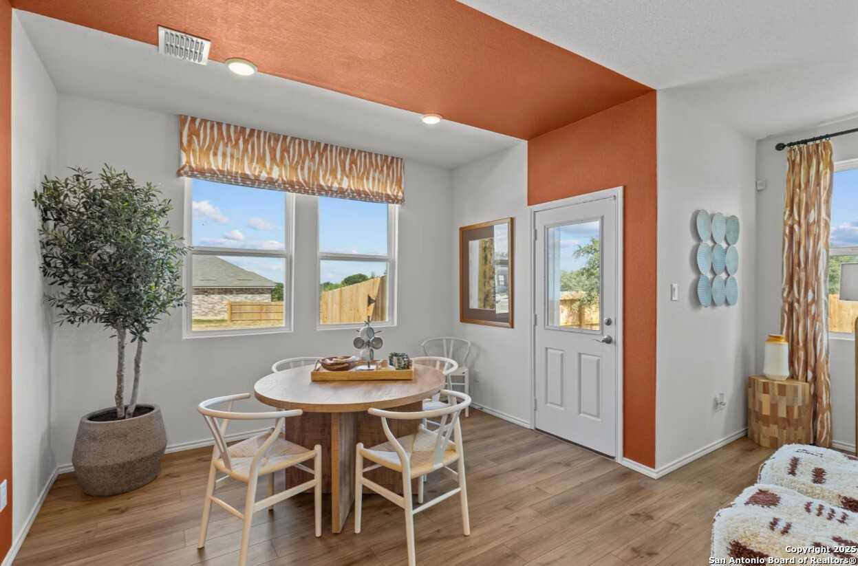 Cozy dining room with round wooden table, white chairs, potted olive tree, and orange accent wall in The Daphne J floor plan, Elmendorf, Texas