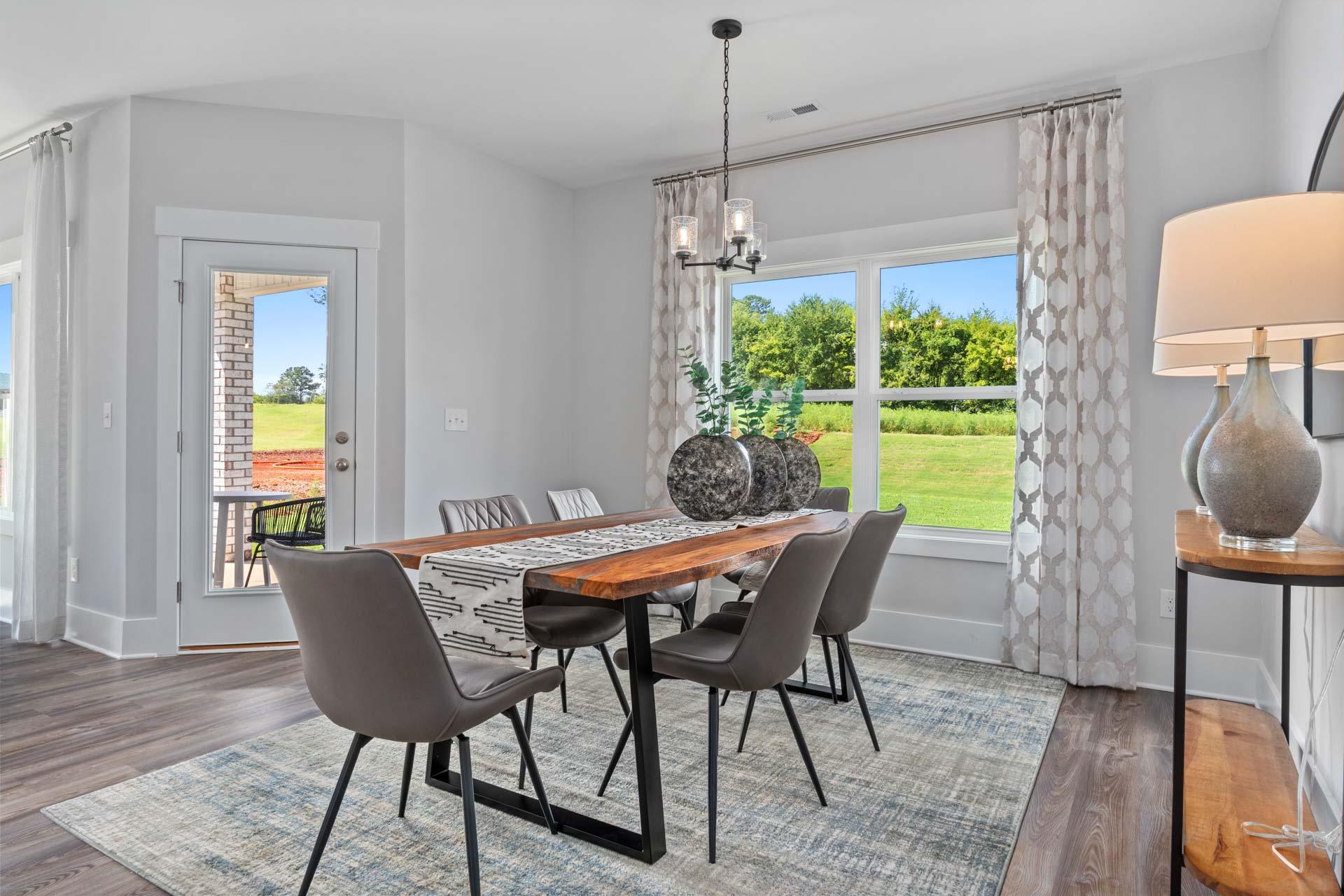 Spacious dining room in Blue Spring Huntsville AL with wooden farmhouse table, gray chairs, chandelier, and lush green backyard views