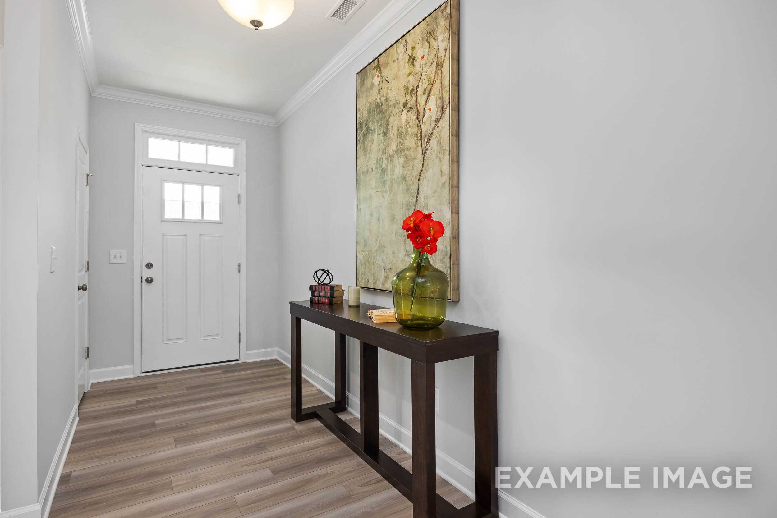 Spacious entryway in The Daphne C home design featuring light wood floors, white door, console table with red flowers in green vase, and abstract wall art
