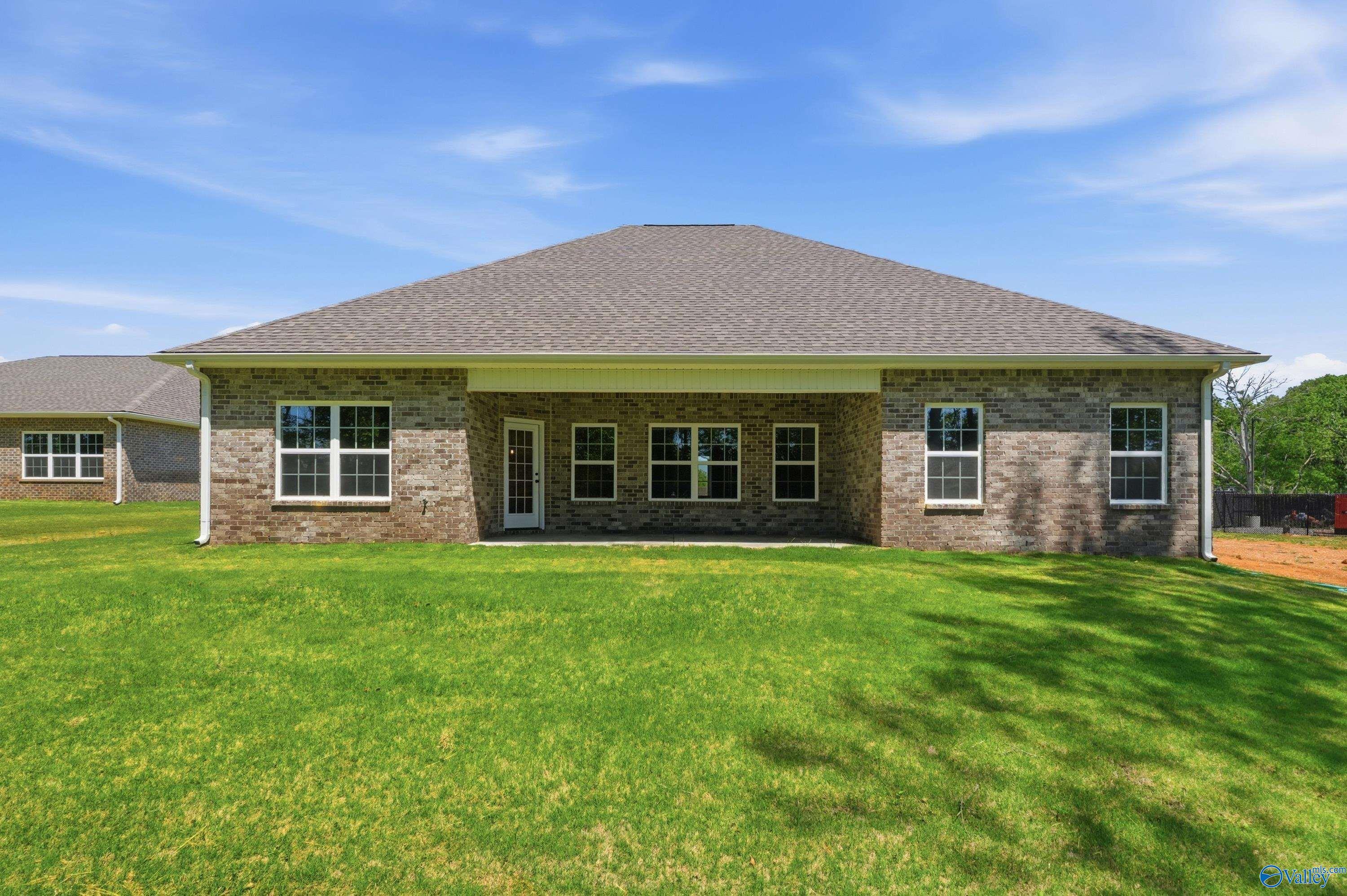 Brick single-story home rear with covered patio, large windows, lush green lawn, and blue sky in The Meadows, Athens, Alabama