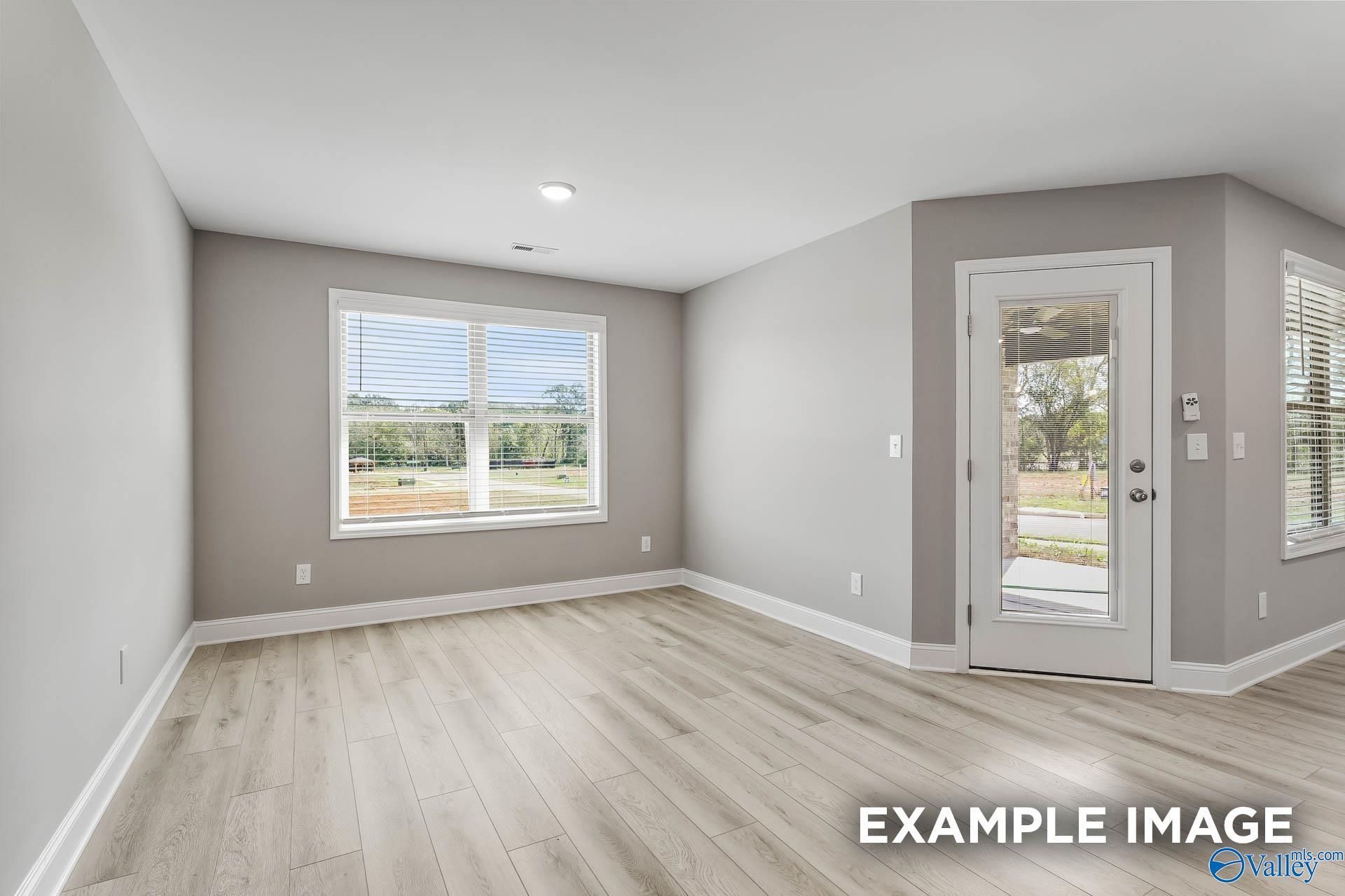 Bright empty living room with gray walls, large windows overlooking trees, sliding glass door, laminate wood floors in The Everett B, Toney, AL