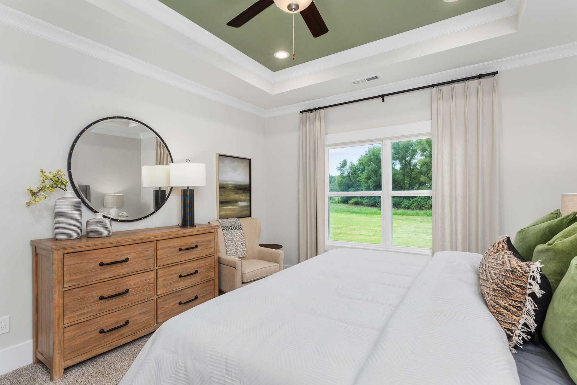 Spacious bedroom at The Meadows in Athens Alabama with wooden dresser, round mirror, white bed, and green window view
