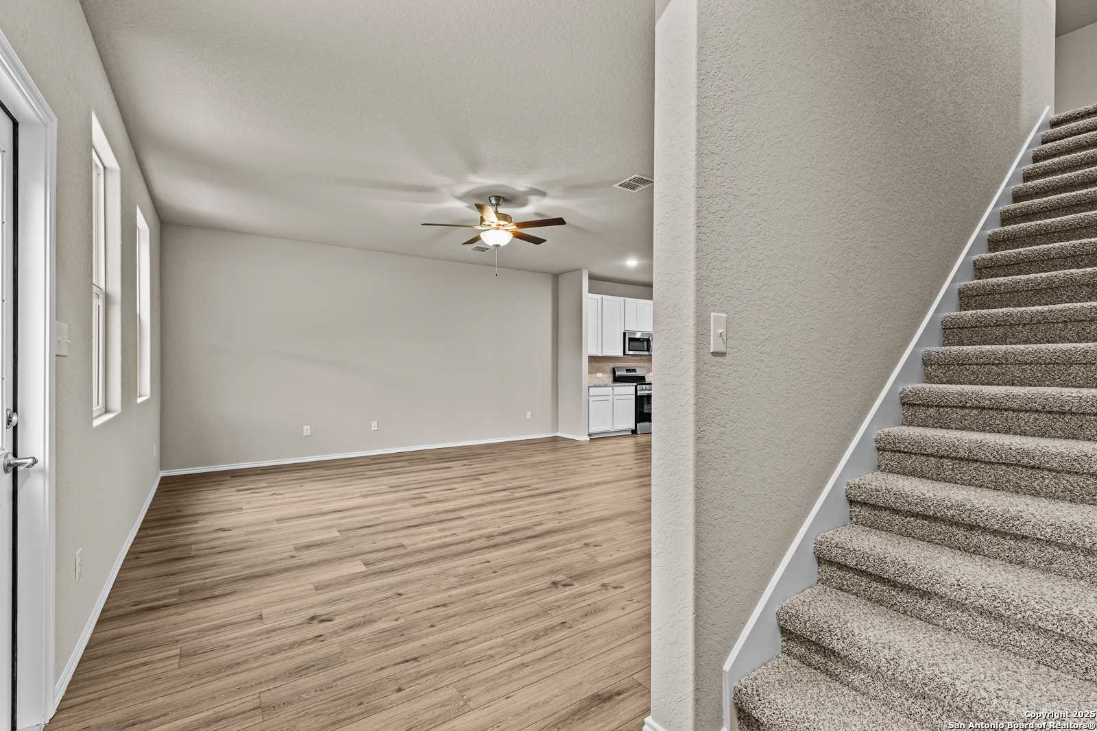 Spacious living area with hardwood floors, ceiling fan, open staircase, and kitchen glimpse in Davidson Homes The Douglas F, San Antonio