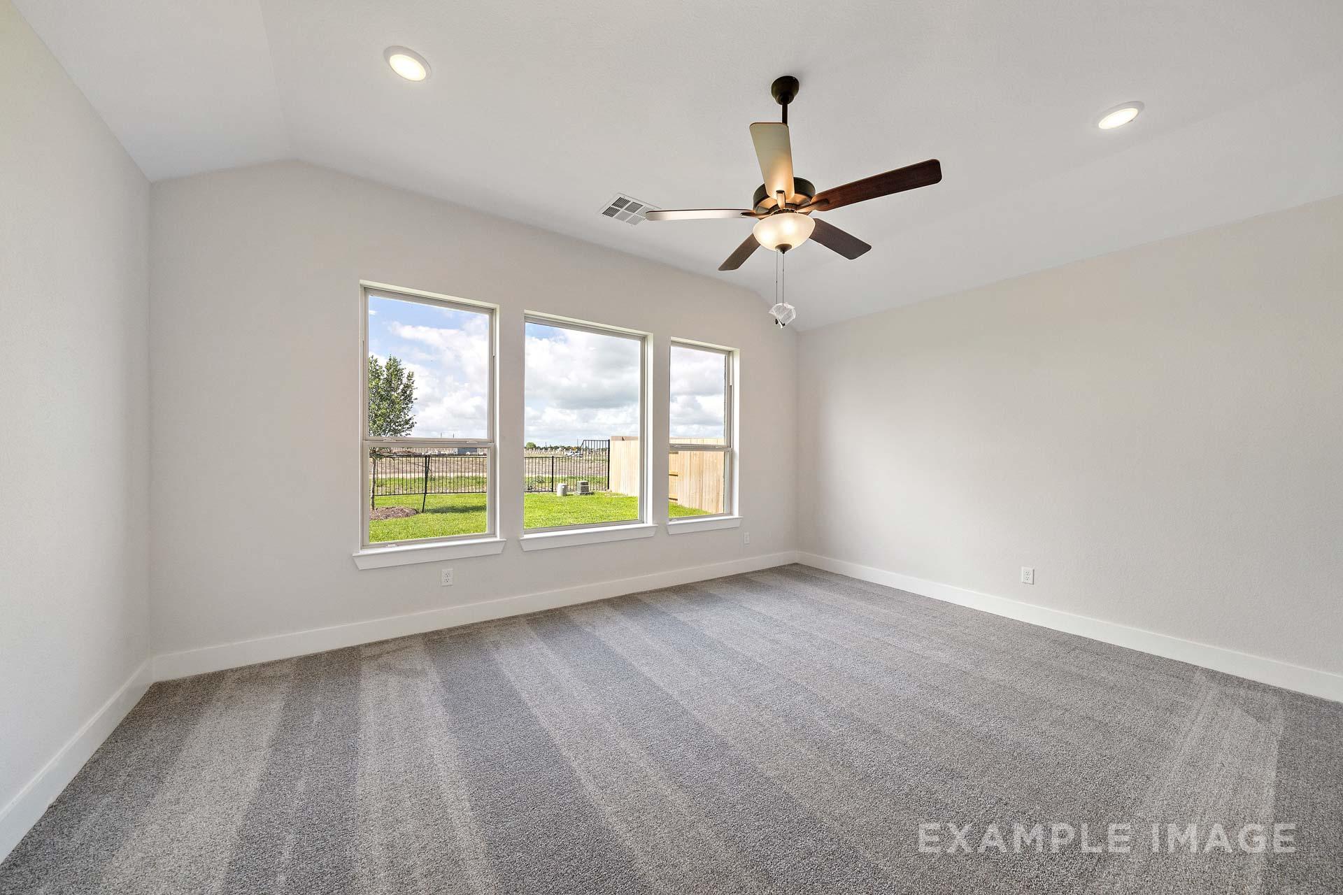 Spacious master bedroom in The Victoria A featuring vaulted ceiling, ceiling fan, triple windows with green backyard view, beige walls, gray carpet